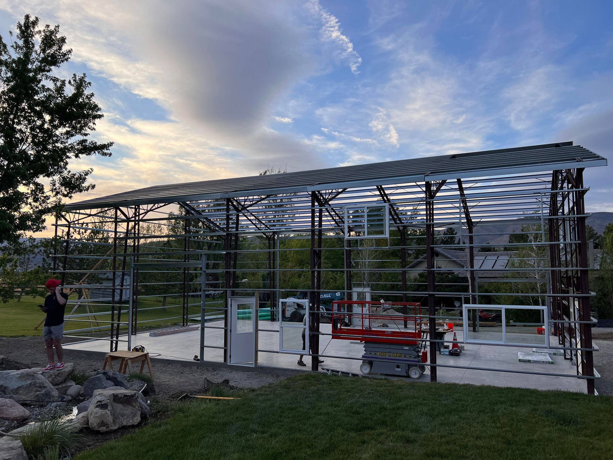 A partially constructed metal frame building, resembling a future shop, sits on a concrete foundation. A person is working near the structure, and construction equipment is visible inside. The sky is cloudy, and trees surround the site at River Bend.