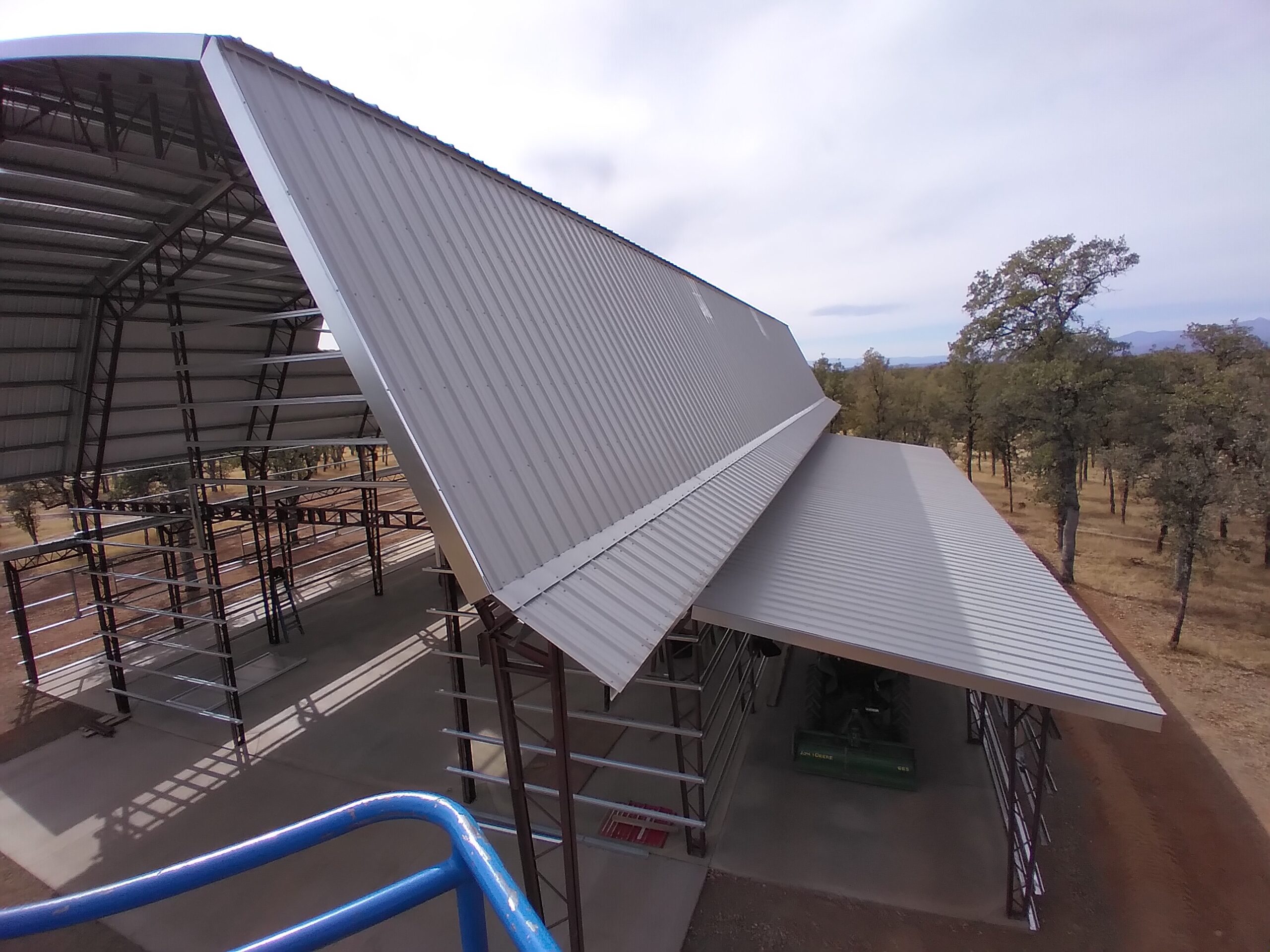 A partially constructed gambrel style metal barnhouse with a sloped roof stands in a rural area. The structure is surrounded by trees and open land. Scaffolding is visible inside, and the sky is overcast.