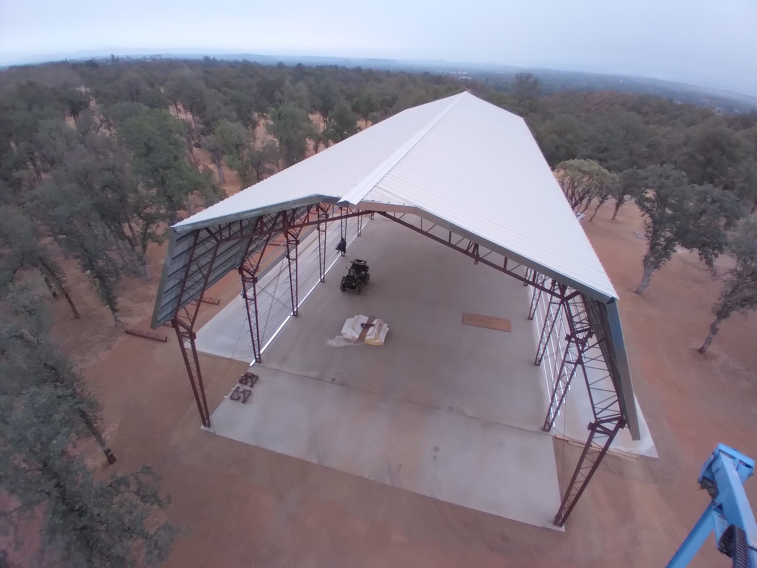 Aerial view of a large open-sided metal barnhouse with a gambrel-style peaked roof, nestled in a clearing surrounded by trees. Beams support the roof, and a few vehicles and equipment are visible on the concrete floor beneath it.