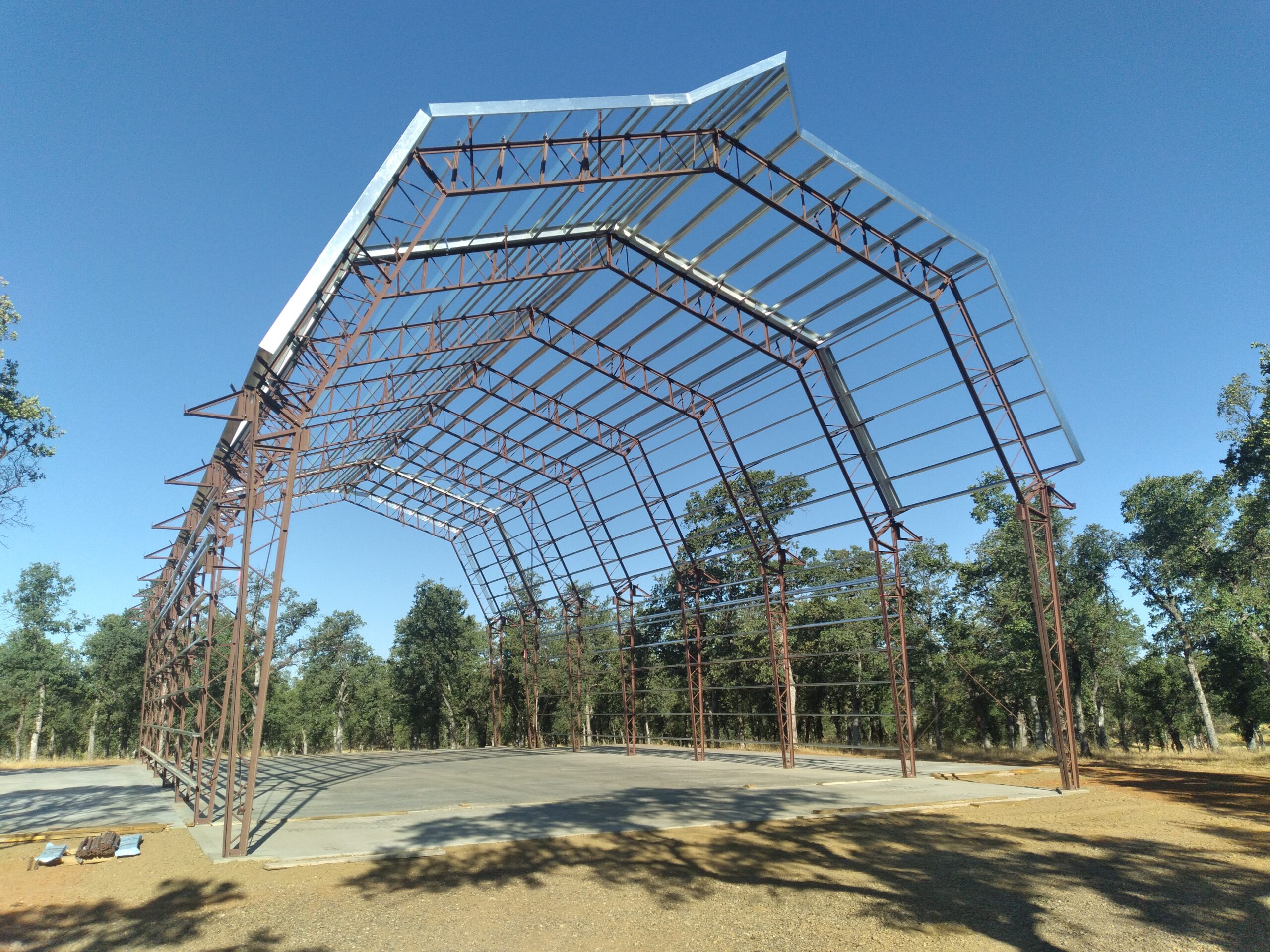 A partially constructed metal frame of a large barnhouse stands on a concrete foundation, surrounded by trees and clear blue sky. The gambrel-roofed structure is open on all sides with visible beams and supports.