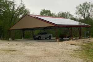 In the tranquil setting of Cedar Hollow, an agricultural metal pavilion with a red roof and solar panels shelters a pickup truck, trailer, and picnic table. The structure rests on a gravel surface, encircled by lush trees.