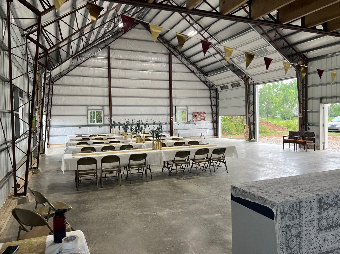 Inside the expansive steel garage, transformed into an elegant venue, long tables draped in white tablecloths showcase gold vases and lush greenery. Folding chairs line both sides as triangular banners dangle from the ceiling. Through an open door, vibrant greenery beckons from outside.