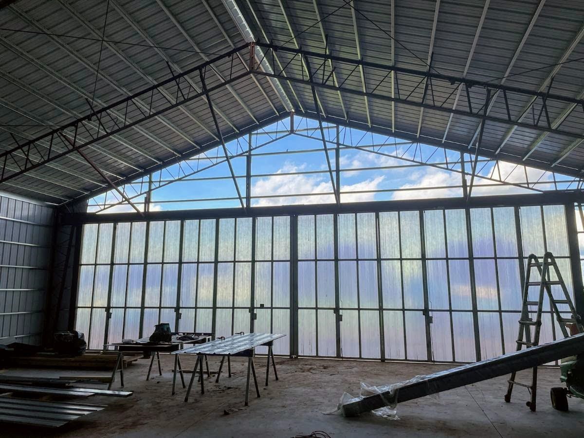 The interior of a spacious industrial building under construction showcases an innovative hangar design, with metal beams and a high ceiling. The semi-transparent wall allows natural light to filter through, revealing a partly cloudy sky. Construction materials and ladders are scattered on the ground.
