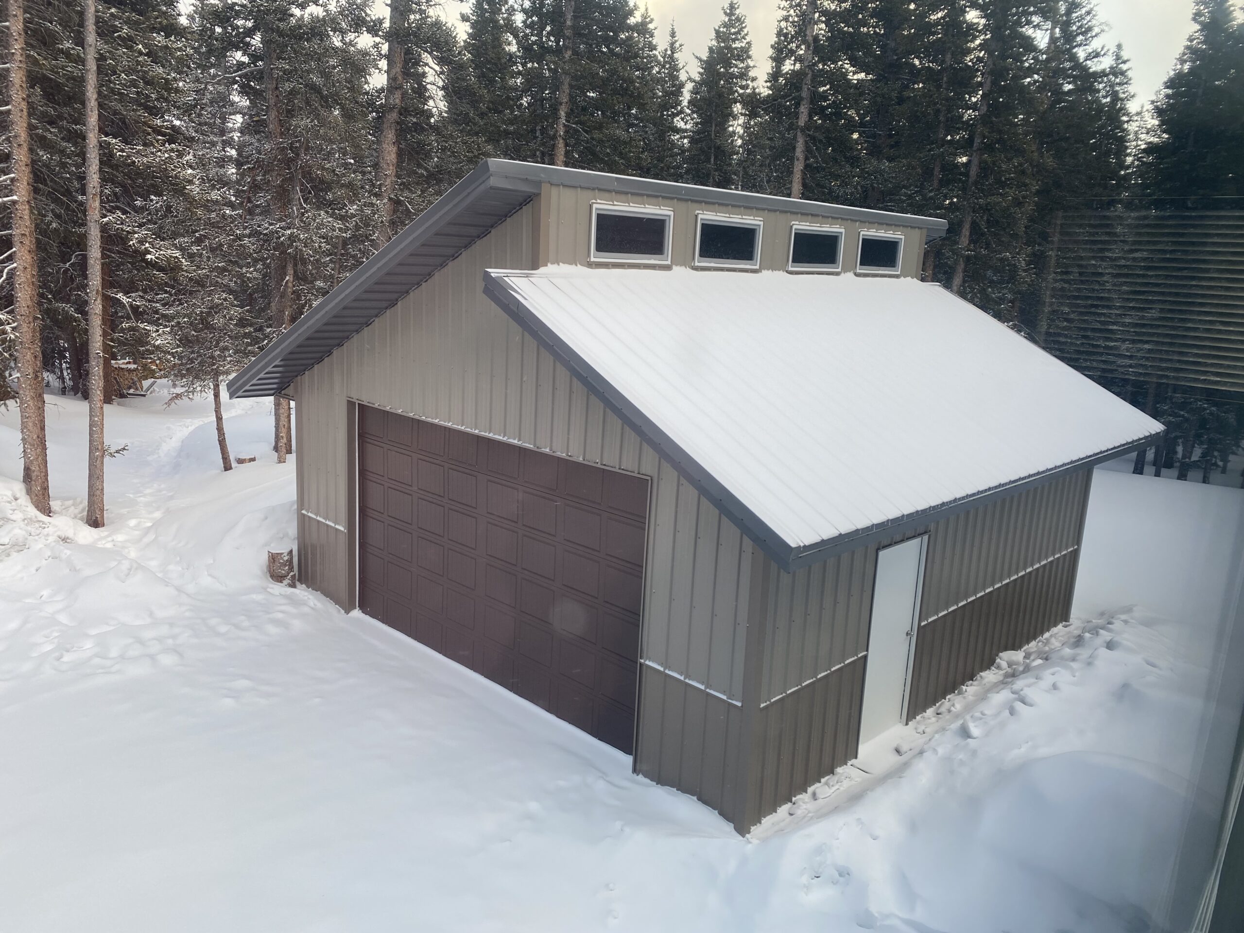 A snow-covered garage with a sturdy metal roof, designed to handle Colorado's hefty snow load, sits amidst a snowy landscape surrounded by tall evergreen trees.
