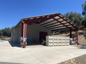 A large open-sided metal building with a red roof and beams stands on a concrete surface, resembling the robust structures found in Belledor vineyards. Stacks of crates and parked equipment are visible inside, while trees and a dirt slope create a picturesque backdrop under the clear blue sky.
