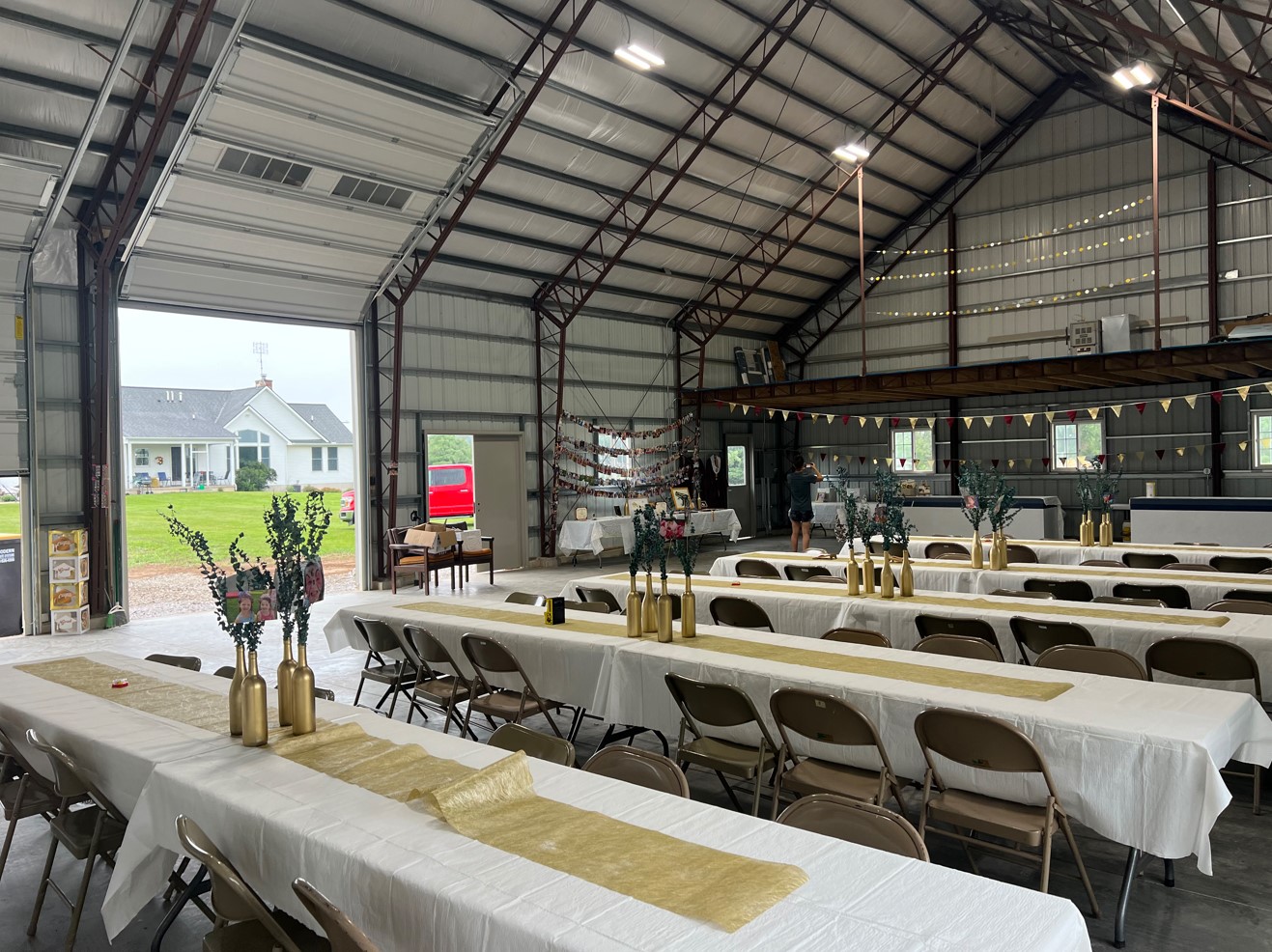 A spacious indoor venue set for an event, featuring long tables with white tablecloths and gold runners. Chairs are neatly arranged, and gold vases with floral arrangements grace the tables. A bar area with bunting adds charm in the background, nestled within a renovated steel garage.