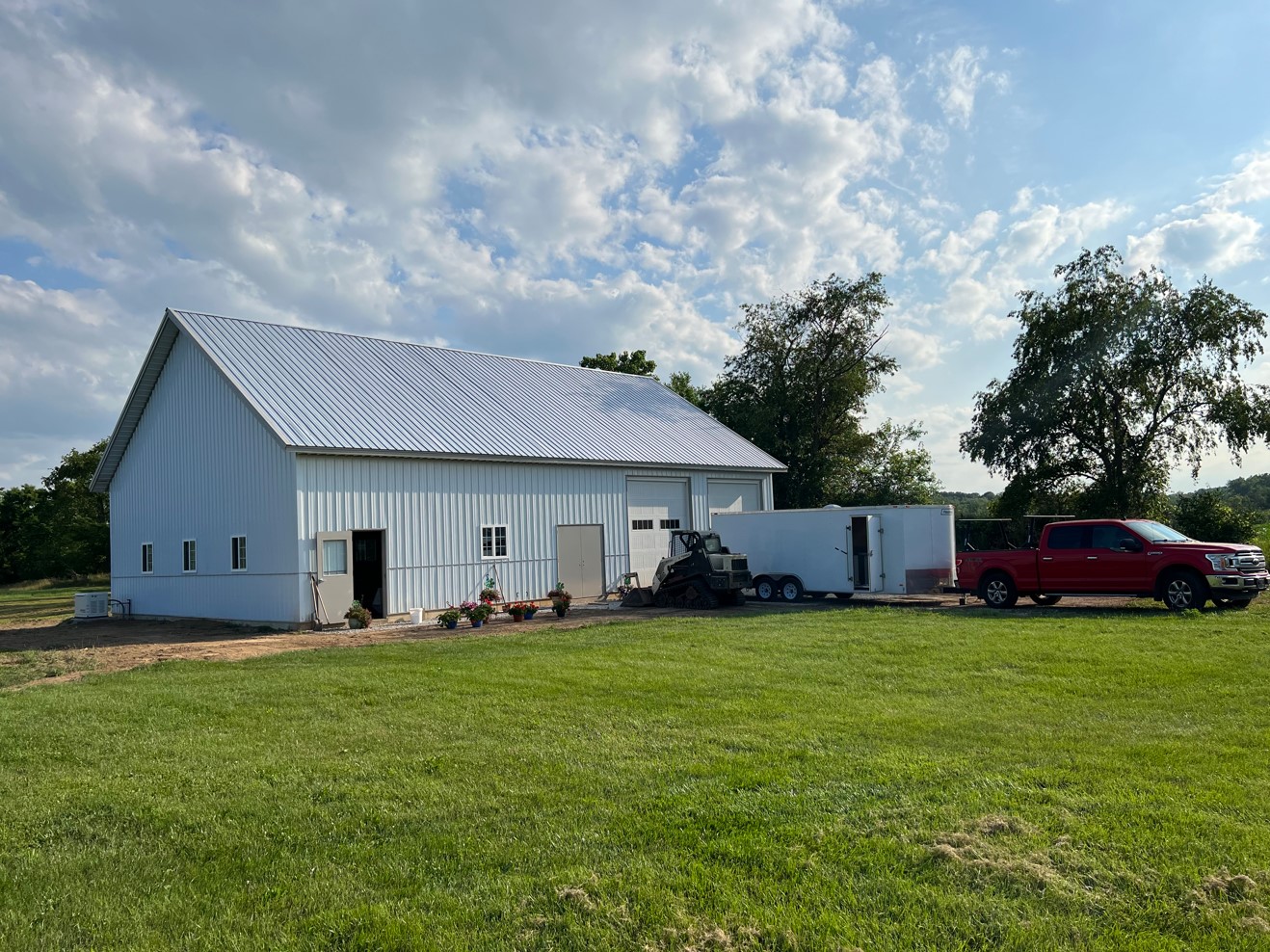 A large white barn with a metal roof stands in a grassy field under a partly cloudy sky, serving as an ideal venue. Nearby, a red truck and trailer are parked alongside equipment and potted plants. In the background, trees complete this picturesque scene.