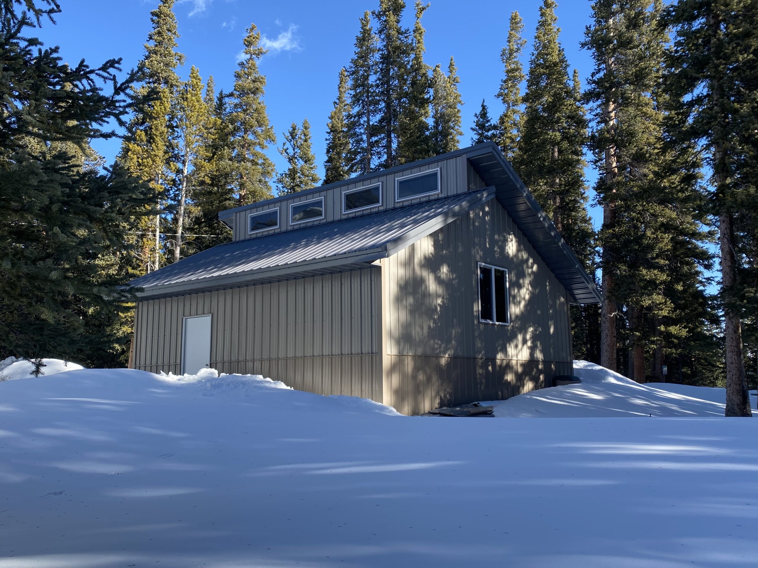 A small, beige cabin with a slanted metal roof stands surrounded by tall evergreen trees in Colorado. Snow covers the ground, and sunlight casts shadows on the snow load, creating a serene, secluded winter scene near the detached garage.