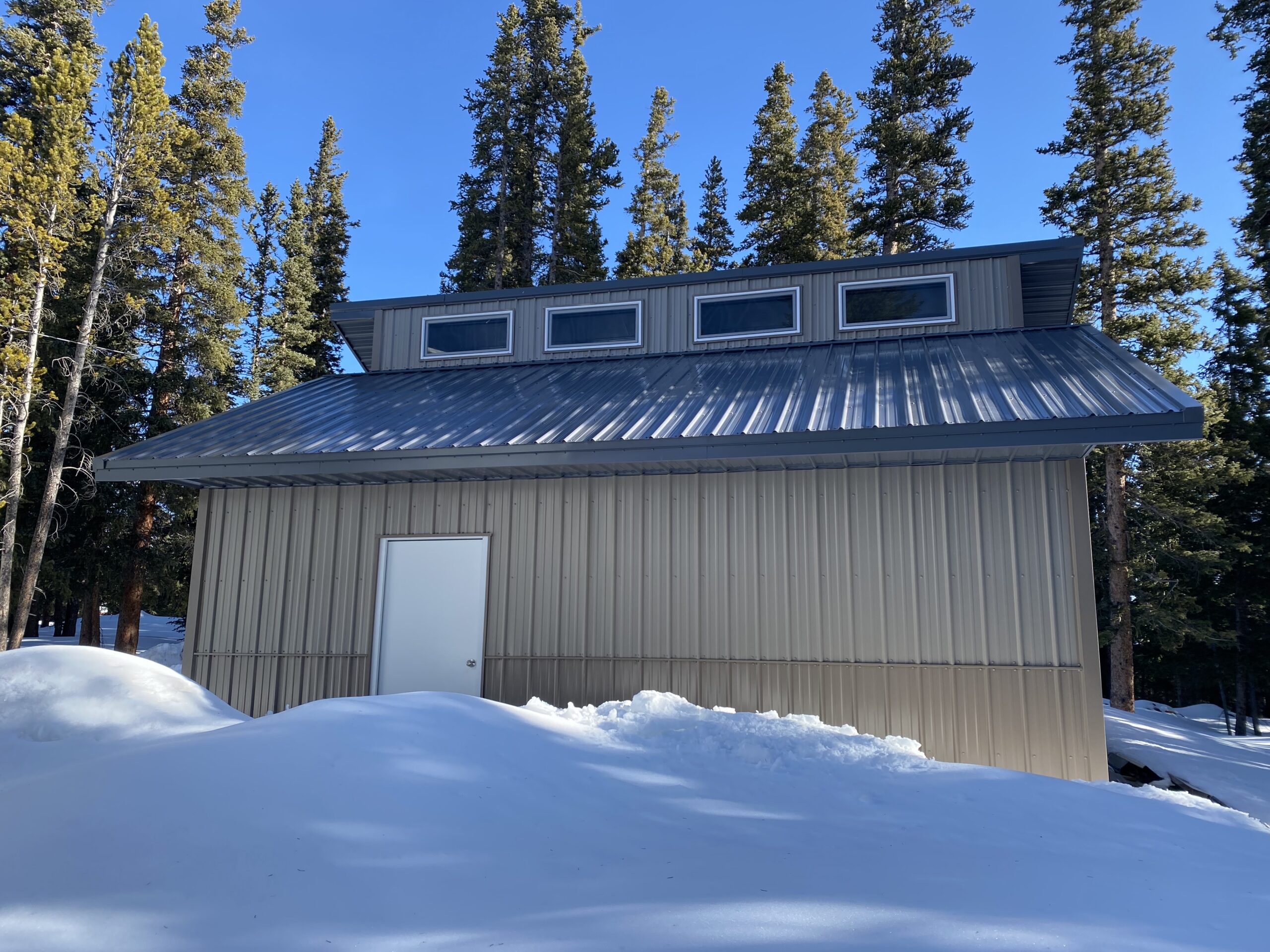A beige metal-sided garage with a sloped roof, designed to withstand the snow load typical of Colorado winters, features three rectangular windows near the top. It stands amidst a snowy landscape, surrounded by tall evergreen trees under a clear blue sky.