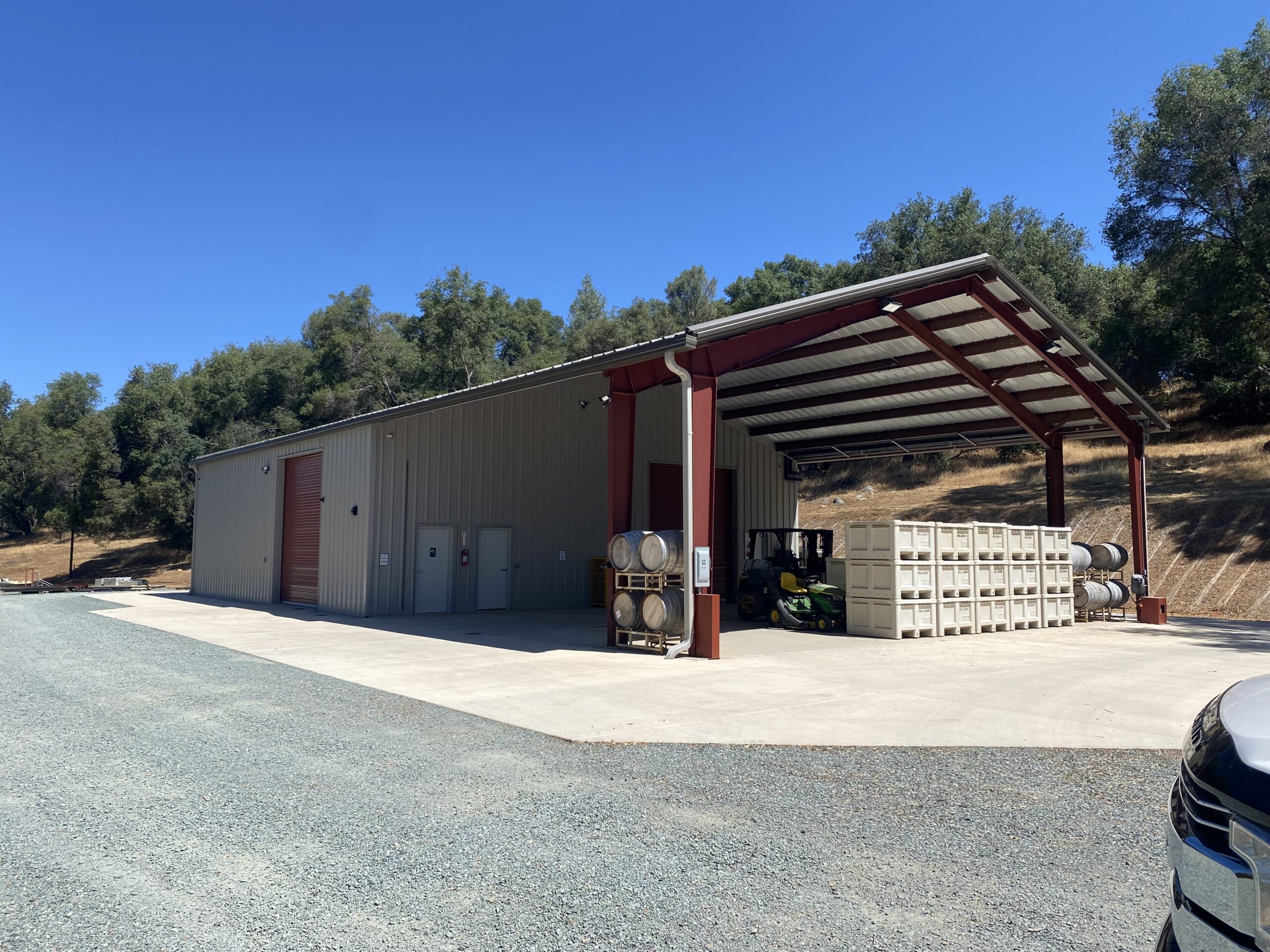A large metal warehouse at Belledor Vineyards boasts an extended open shelter, housing barrels and crates. A tractor is parked beneath the shelter. The building is encircled by gravel, trees, and a clear blue sky, enhancing this vineyard's rustic charm.