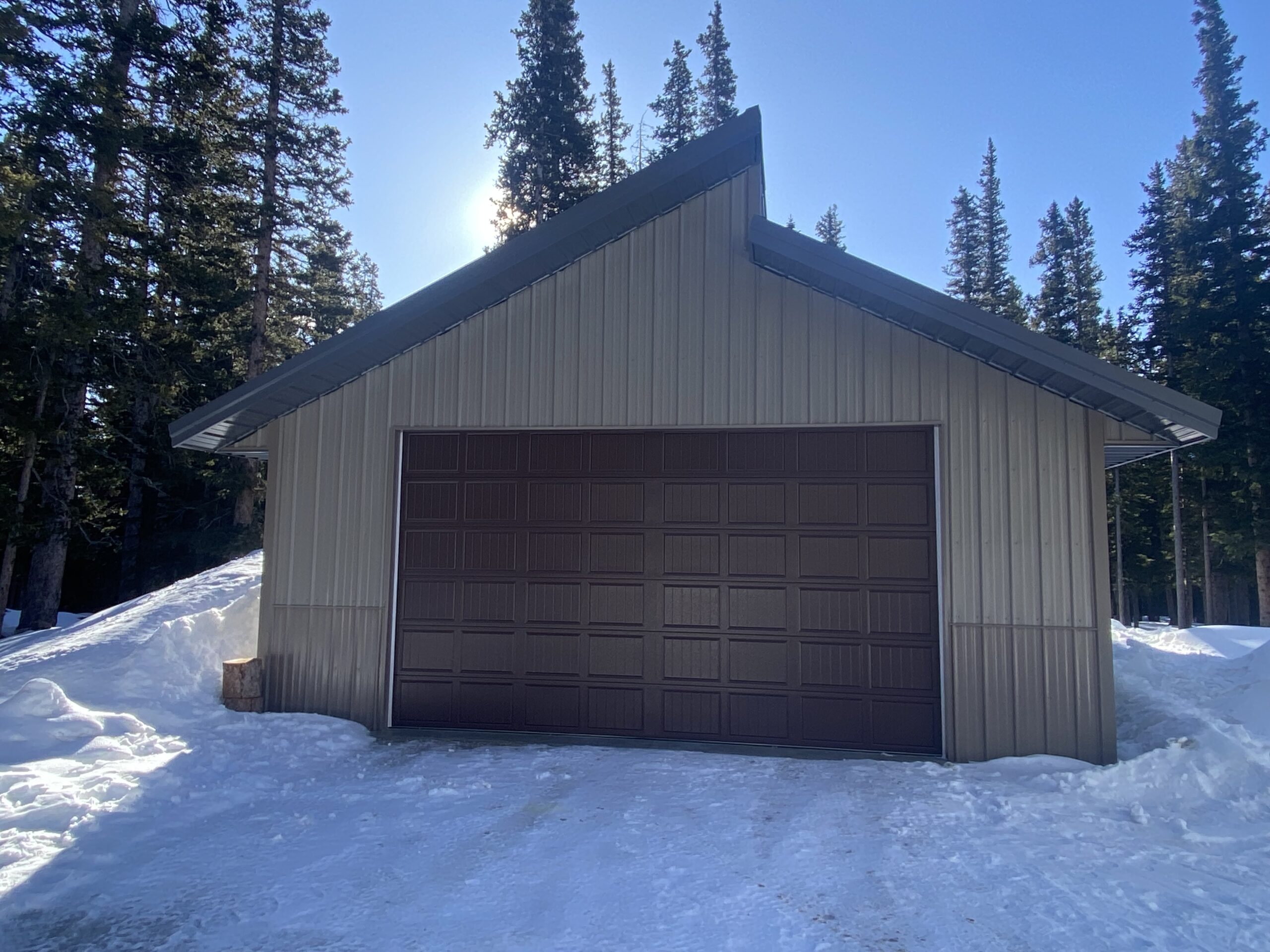 A metal garage with a dark brown door is enveloped by snow, managing its snow load efficiently. Towering pine trees rise in the background under Colorado's clear blue sky, while sunlight casts a gentle glow from behind the structure.