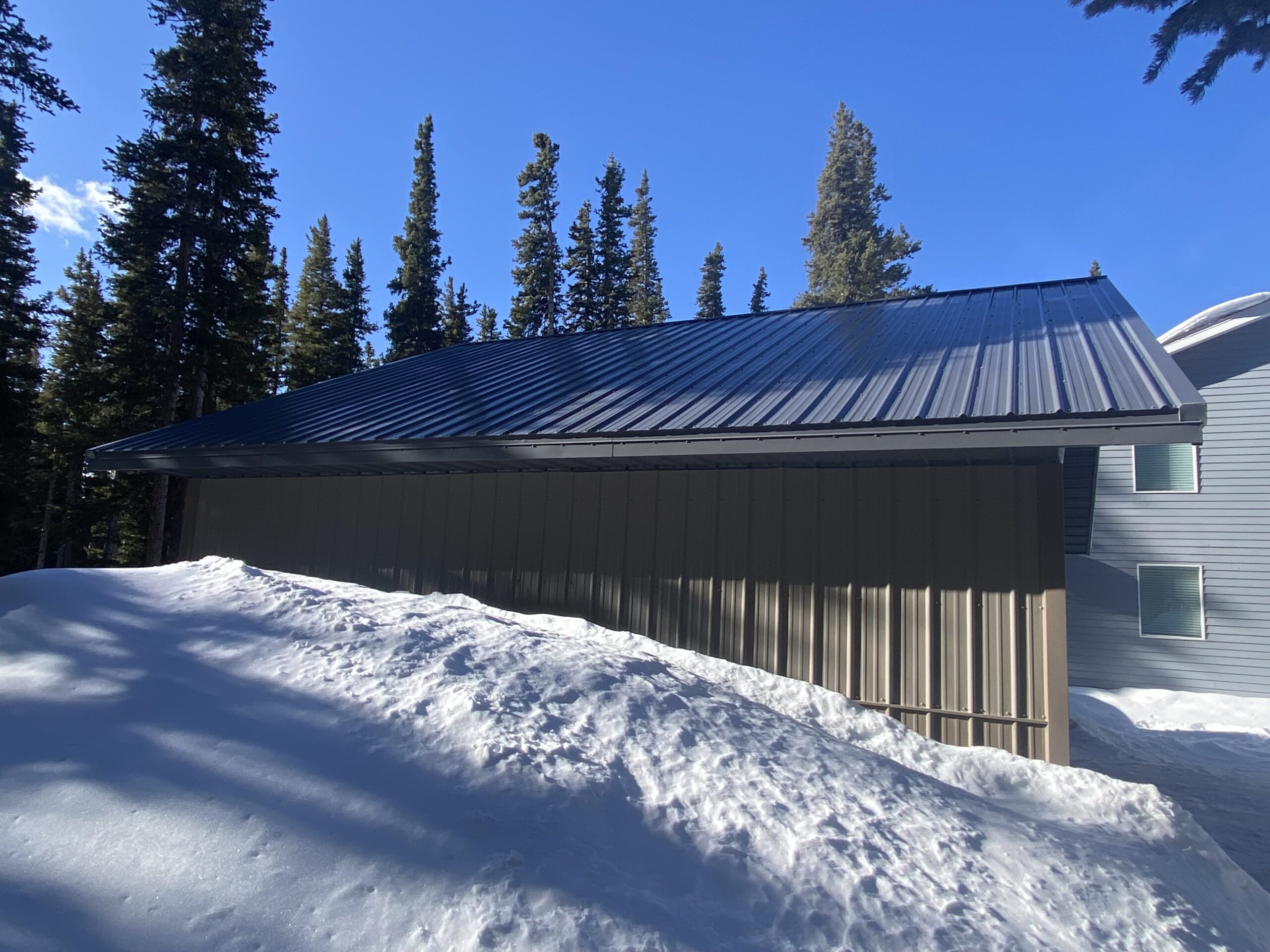 A modern building with a dark metal roof sits partially covered in snow, designed to withstand Colorado's substantial snow load. Tall evergreen trees surround the structure under a bright blue sky, and snow piles are visible in the foreground, hinting at its adjacent garage.