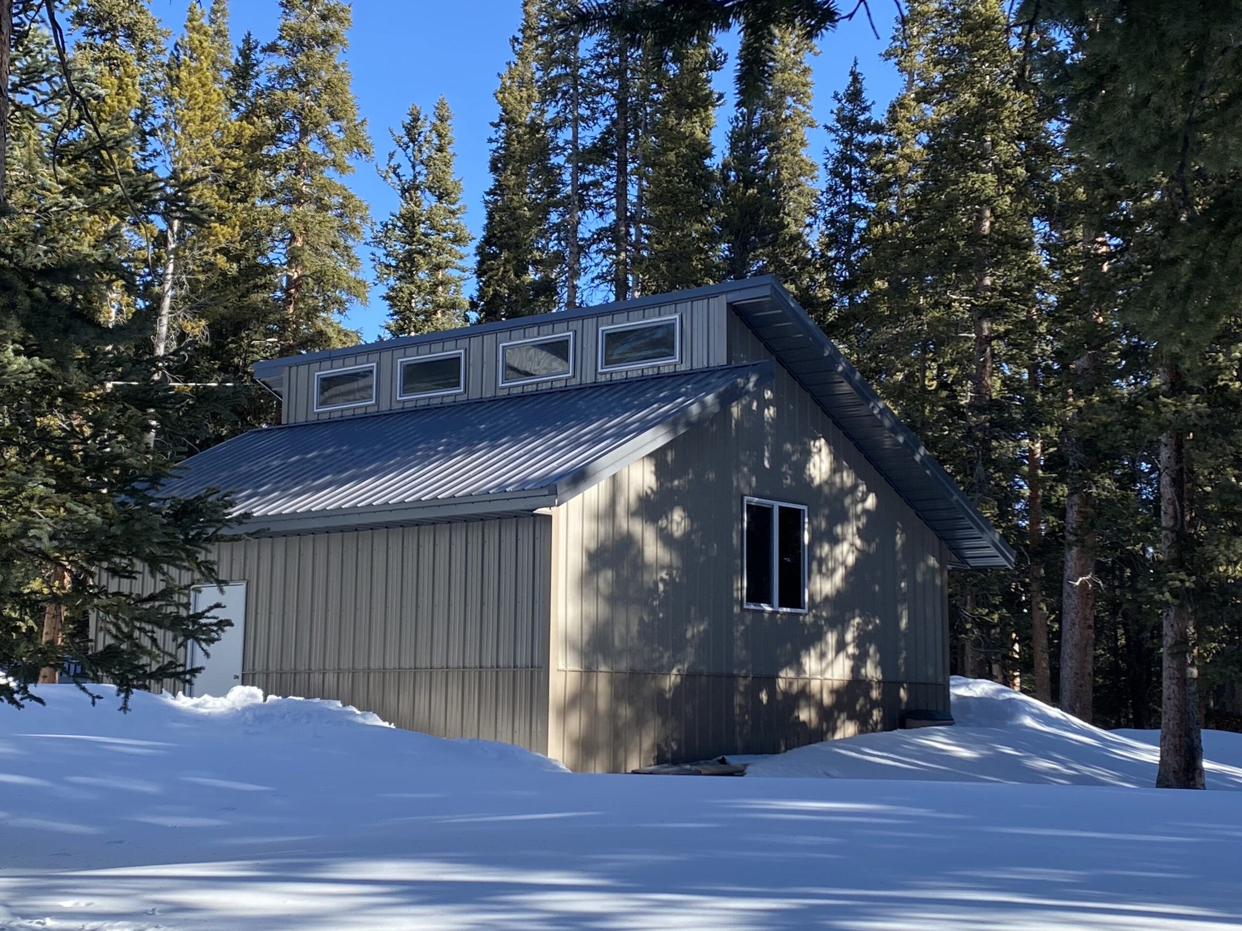 A small, beige cabin with a metal roof stands in Colorado's vast wilderness, surrounded by tall pine trees. Sunlight filters through the branches, casting shadows on the snow-covered ground and highlighting the sturdy construction designed to withstand heavy snow loads. The sky is clear and blue.