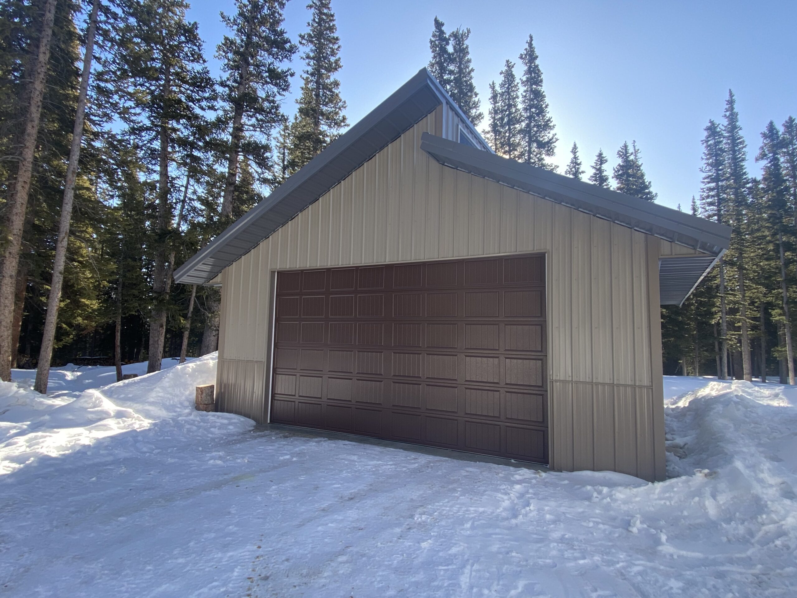 A large metal garage designed to withstand a heavy snow load stands with a brown door in a snowy, wooded area of Colorado. Tall evergreen trees envelop the structure, and the clear blue sky stretches above.