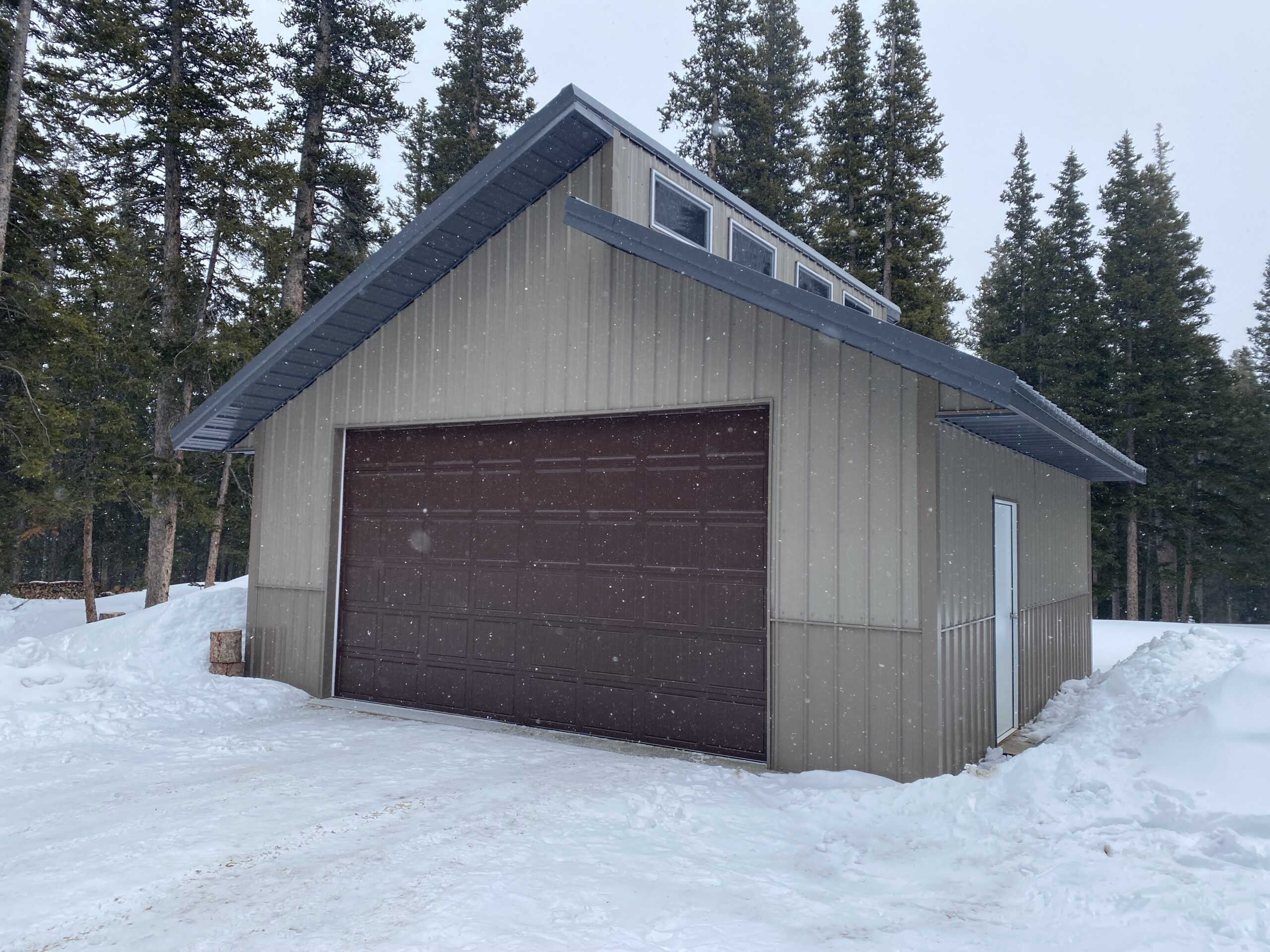 A gray metal garage with a dark brown door is nestled in a snowy Colorado landscape. Evergreen trees surround the structure, designed to withstand heavy snow loads, as flakes gently fall from the overcast sky.