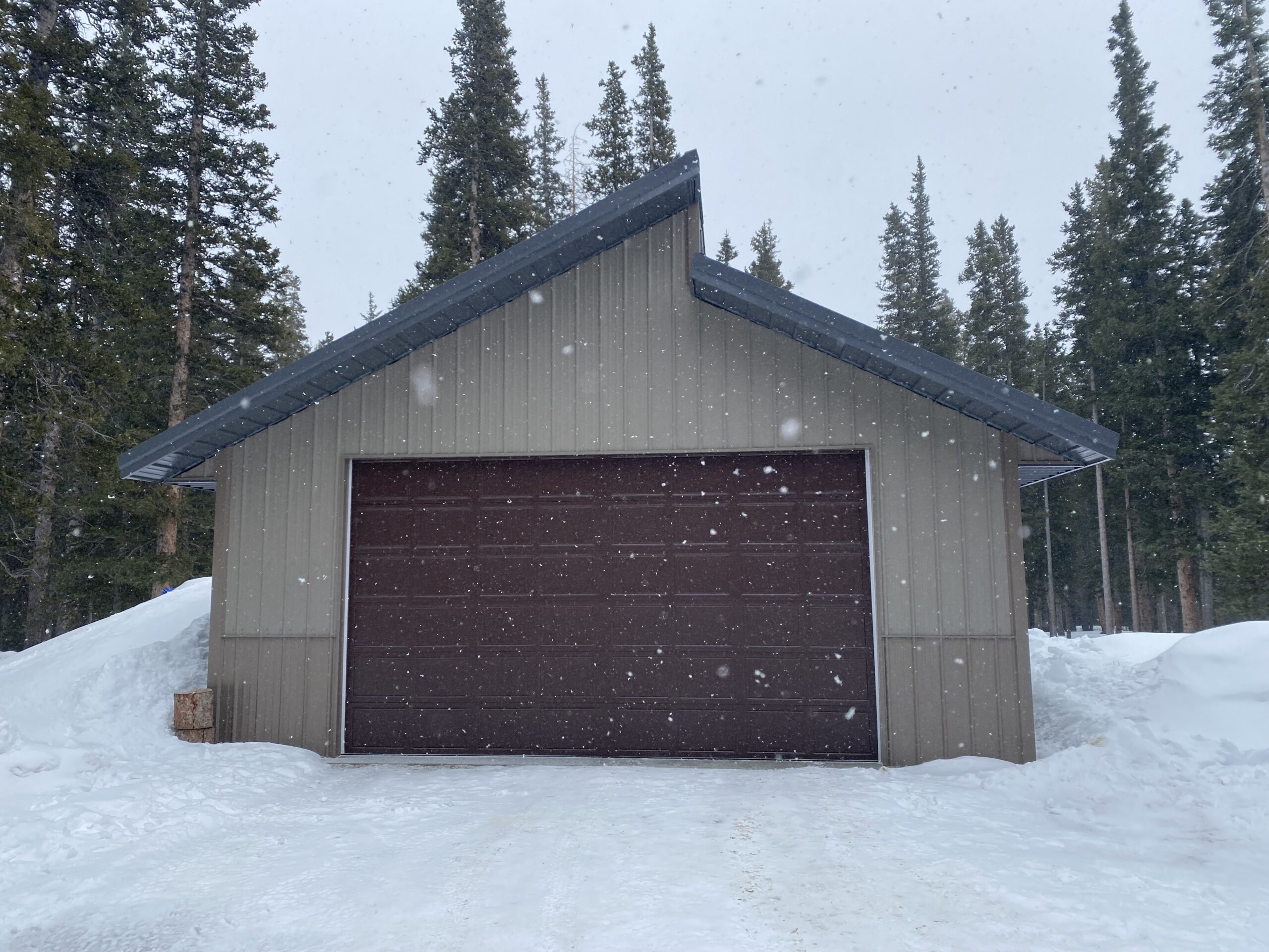 A brown, closed garage in Colorado is nestled amidst snow and pine trees, with delicate snowflakes descending from a gray sky. The building features a sloped, dark roof designed to withstand the region's heavy snow load, perfectly complementing the serene wintry landscape.