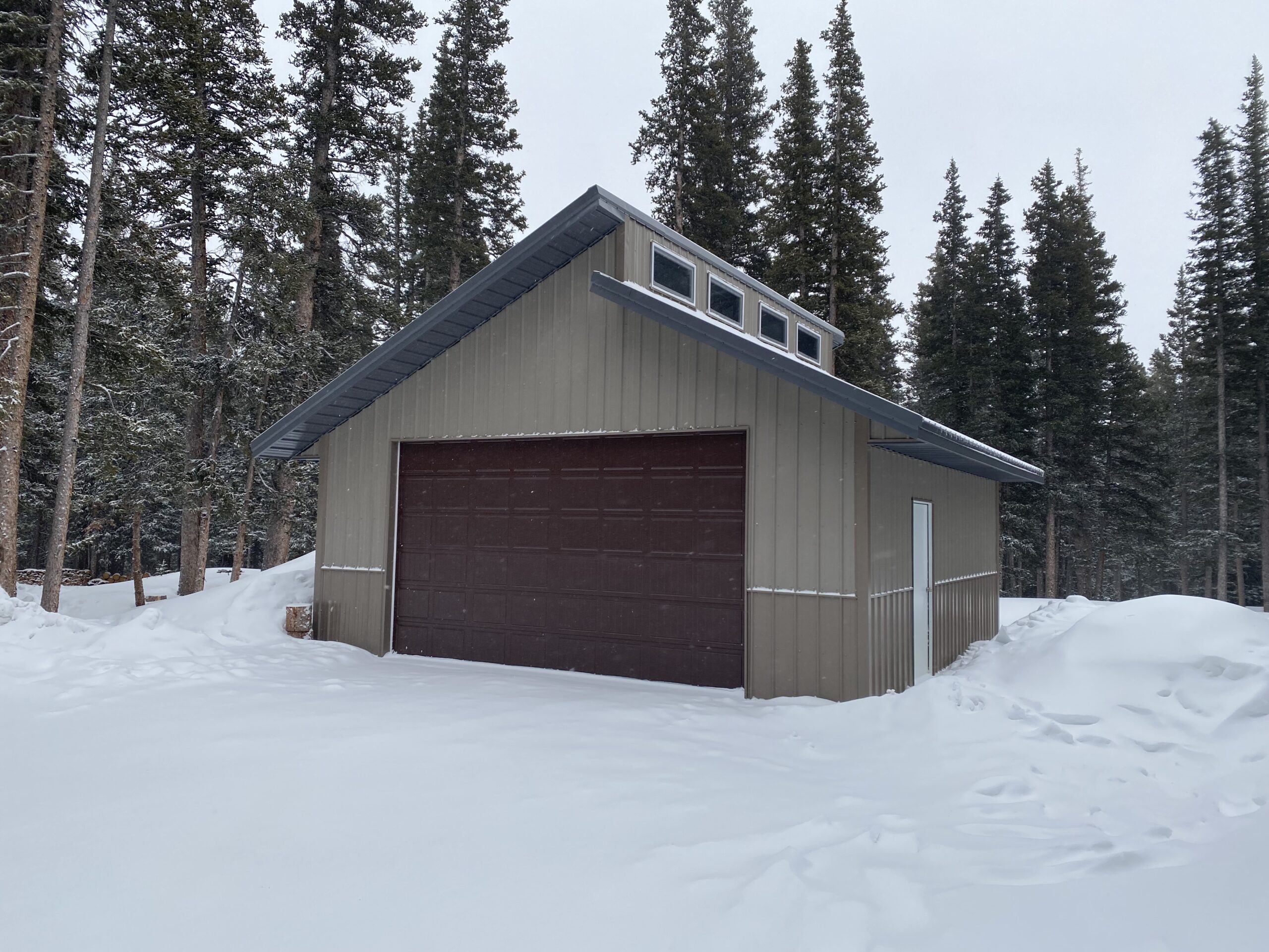 A brown garage with a green roof stands proudly amidst a snowy Colorado landscape, surrounded by tall evergreen trees. The ground is blanketed in snow, and the overcast sky hints at the increasing snow load on this cold, wintry day.