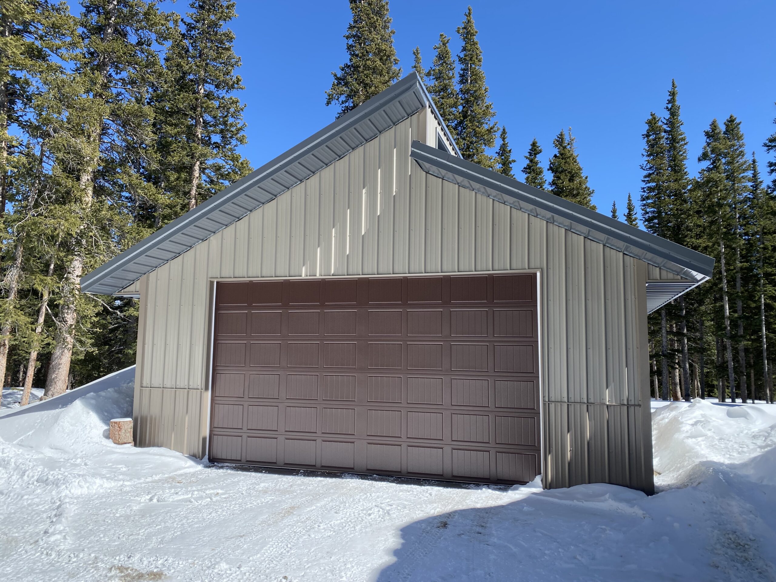 A brown garage in Colorado stands resilient amid a snowy landscape, framed by tall evergreen trees under a clear blue sky, thoughtfully designed to withstand any snow load.