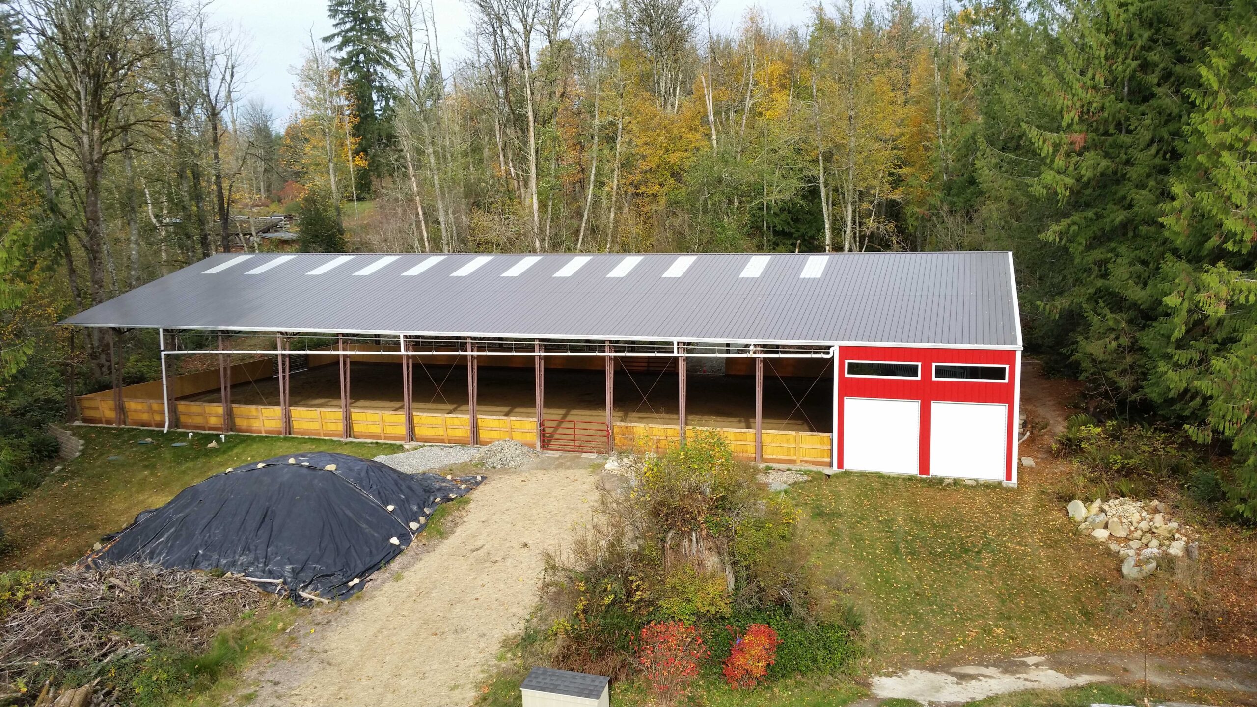 A large red and white barn with a metal roof set against a backdrop of tall trees. There is a dirt path leading to the barn and a covered pile of materials in front. The trees have autumn foliage in shades of yellow and orange.