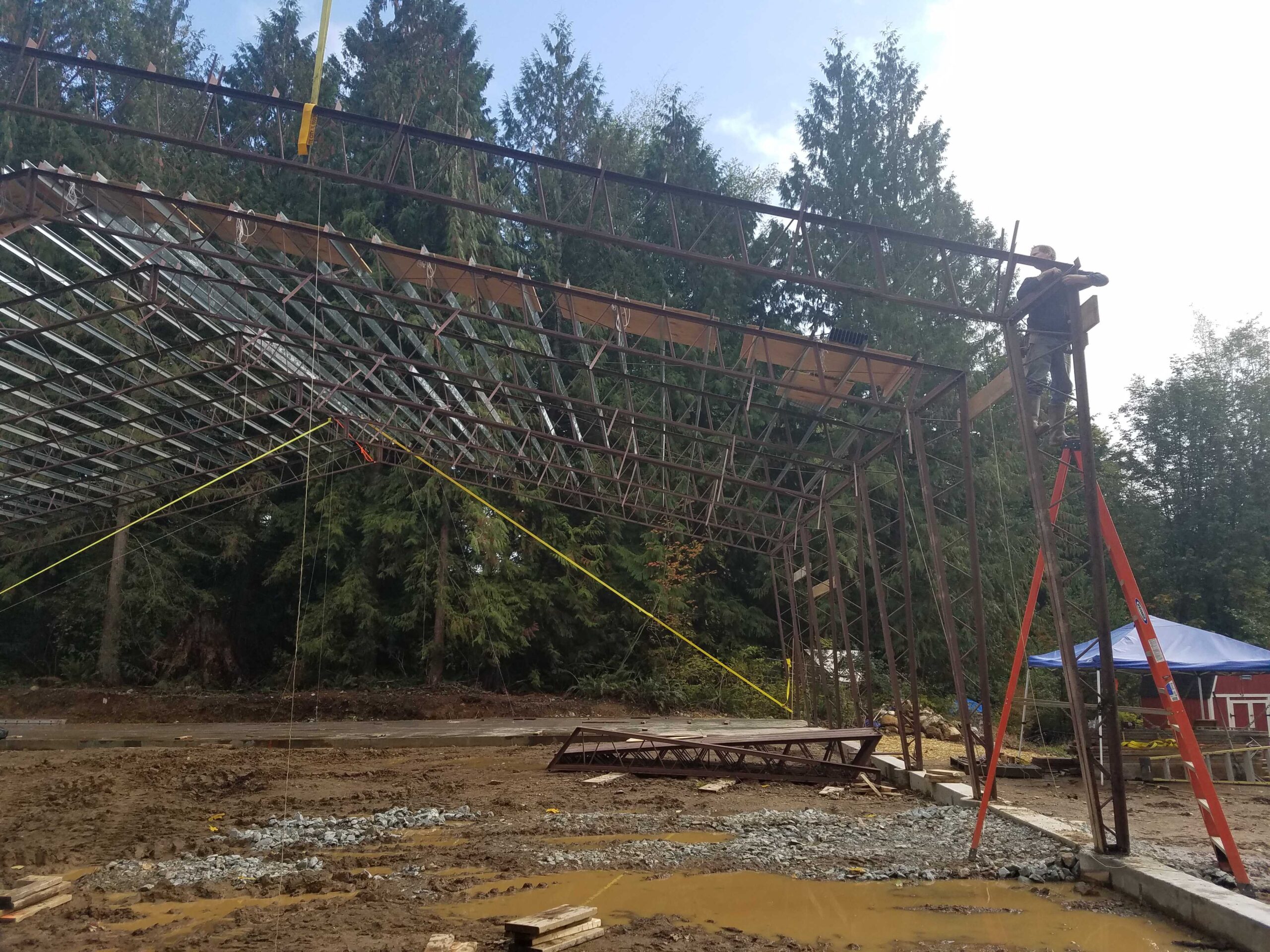 Construction site with a partially built metal structure. A worker stands on scaffolding, assembling beams. Trees are in the background, and the ground is muddy with scattered materials. A blue tent is visible to the right.