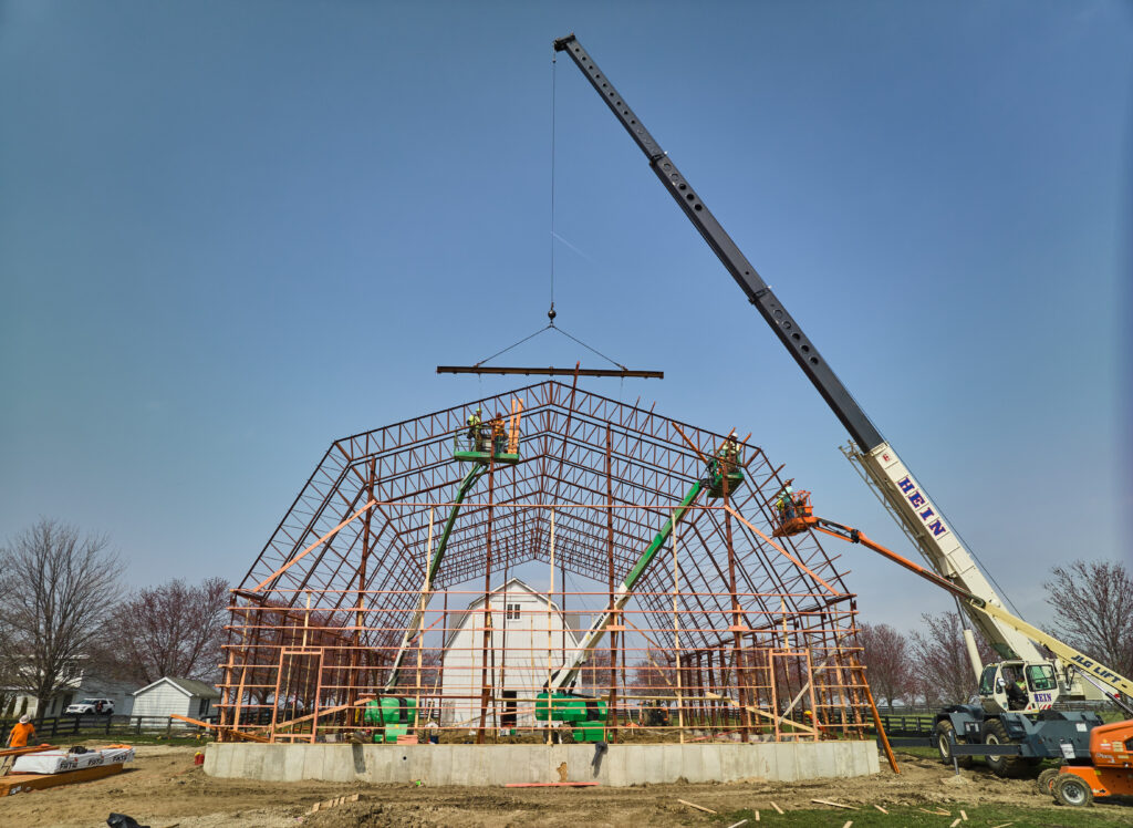 A large crane is constructing a barn's metal framework on a clear day. Two smaller lifts are positioned inside, with workers installing beams. Trees and buildings can be seen in the background. The structure stands on a concrete foundation.