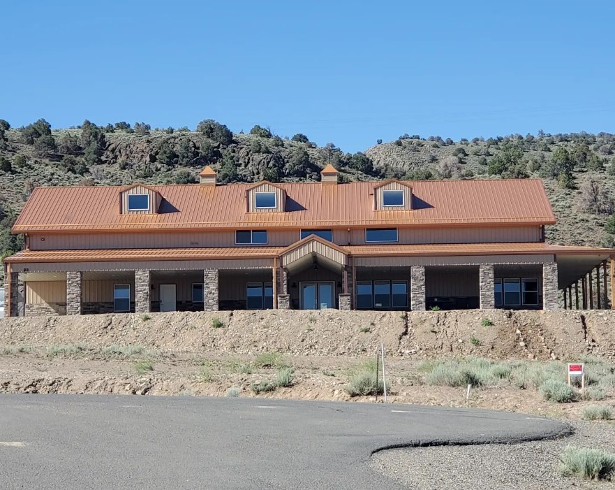 A large, modern barndominium with a sienna roof and stone accents stands on a dirt hill. It features multiple windows and glass doors, set against a backdrop of trees and rocky terrain under a clear blue sky.