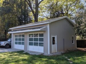 In the backyard, a metal garage with two large glass doors stands proudly in a grassy yard. A silver SUV is parked beside it. Tall trees with lush green foliage surround the structure, as sunlight filters beautifully through the canopy.