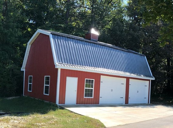 A red barn-style gambrel garage with a silver metal roof and two double garage doors stands on a concrete driveway. Surrounded by trees, this charming structure features a white door and several windows on its side, making it an idyllic garage shop setting.