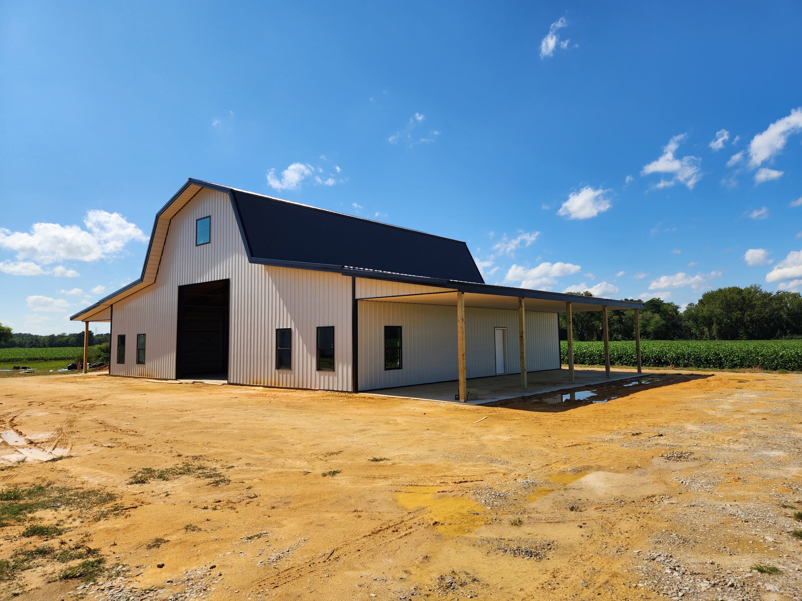 A large white barn with a blue gambrel roof stands under a clear blue sky. The barn features open and closed doors and a lean-to supported by wooden posts. It is surrounded by a dirt surface, with green fields and trees in the background.