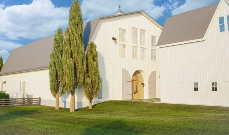 A charming gambrel-roofed barn with a wooden arched door, surrounded by tall, slender trees, is nestled on a grassy landscape under a partly cloudy sky—a perfect wedding venue.