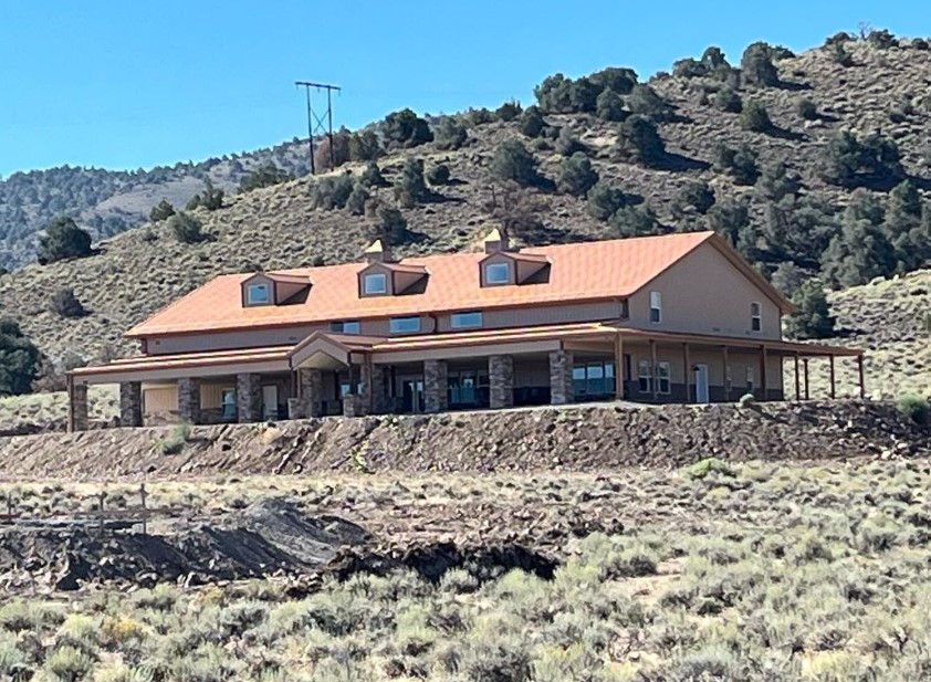 A spacious, two-story barndominium with a sienna roof sits on a sloped, rocky terrain surrounded by desert-like vegetation. The building features multiple windows and a porch. Hills and sparse trees are visible in the background under a clear blue sky.