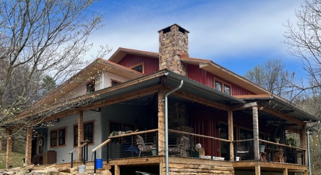 A rustic, asymmetrical two-story house with a wraparound porch, red and gray exterior, and a stone chimney. This charming lakehouse is surrounded by leafless trees under a blue sky with wispy clouds.