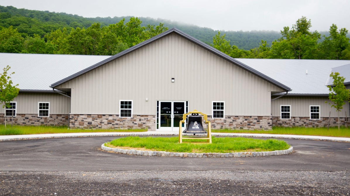 A large, beige building with a stone facade and a metal roof sits against a backdrop of green, tree-covered hills. A riding arena complements the scene as a roundabout with a sign graces the entrance, while a cloudy sky looms above.
