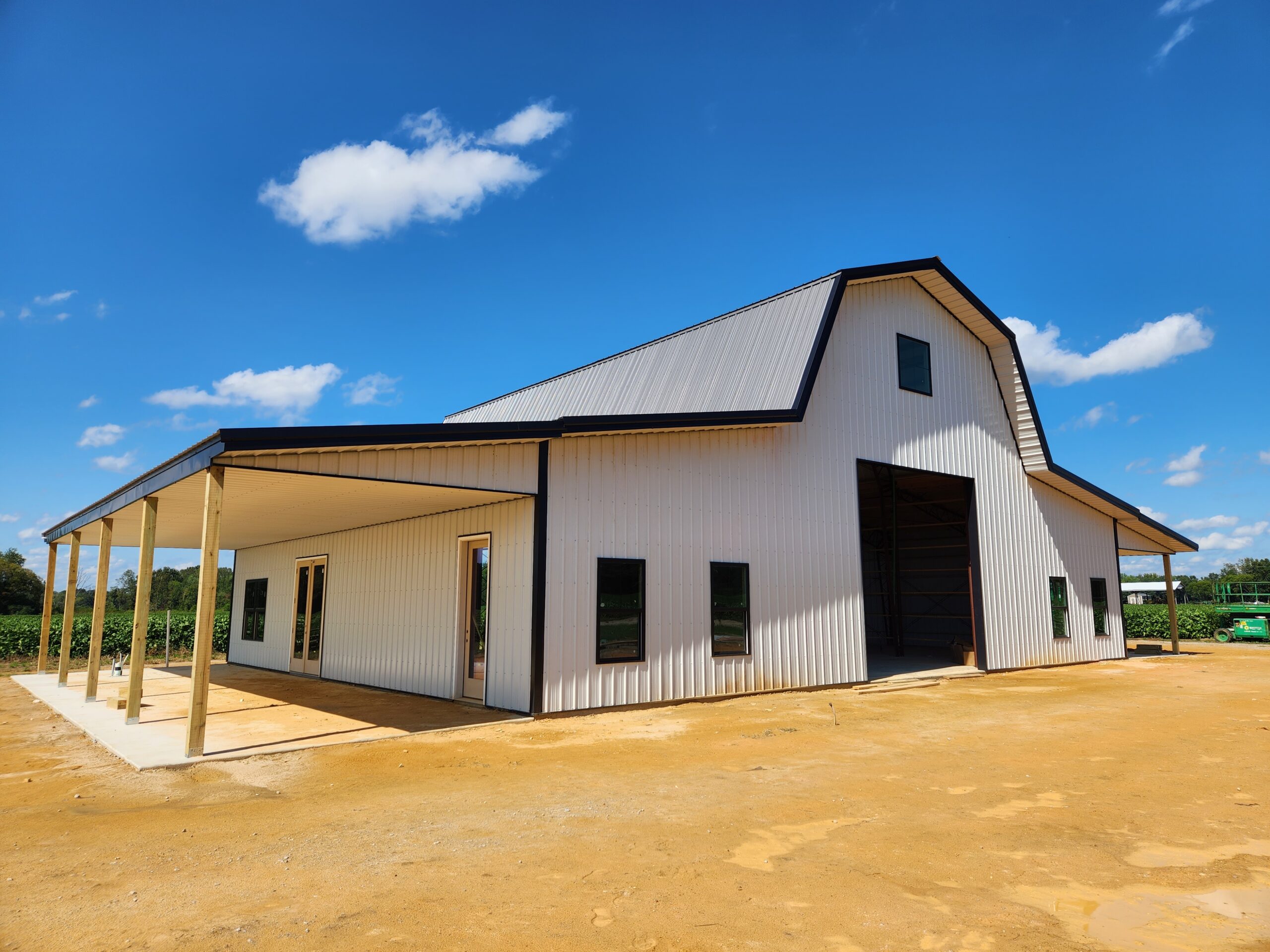 A newly constructed white barn with a dark roof and gambrel design stands on a clear day with a few clouds. The building features a large open entrance and covered area supported by wooden posts. The sandy ground leads to fields visible in the background.