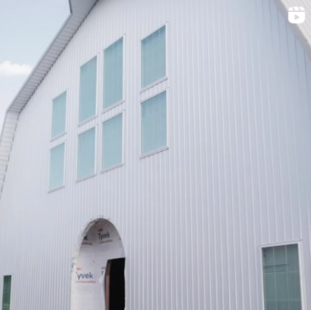 A large, white, modern barn structure with tall vertical siding and multiple rectangular windows stands proudly. The wide arched entrance is under construction, hinting at its future as a stylish wedding venue. Building materials are visible at the lower part, awaiting their final purpose.