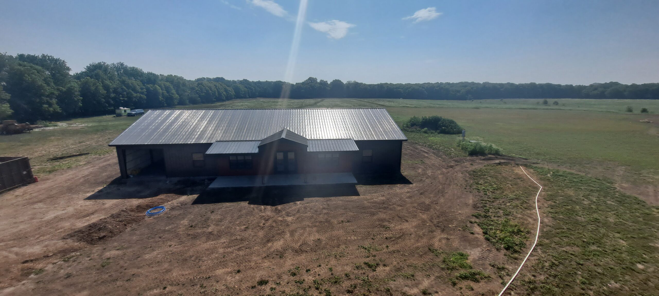 A large metal-roofed barndo sits in the middle of a clearing, surrounded by expansive green fields and a line of trees in the background, under a bright blue sky with scattered clouds at Reece Ranch.