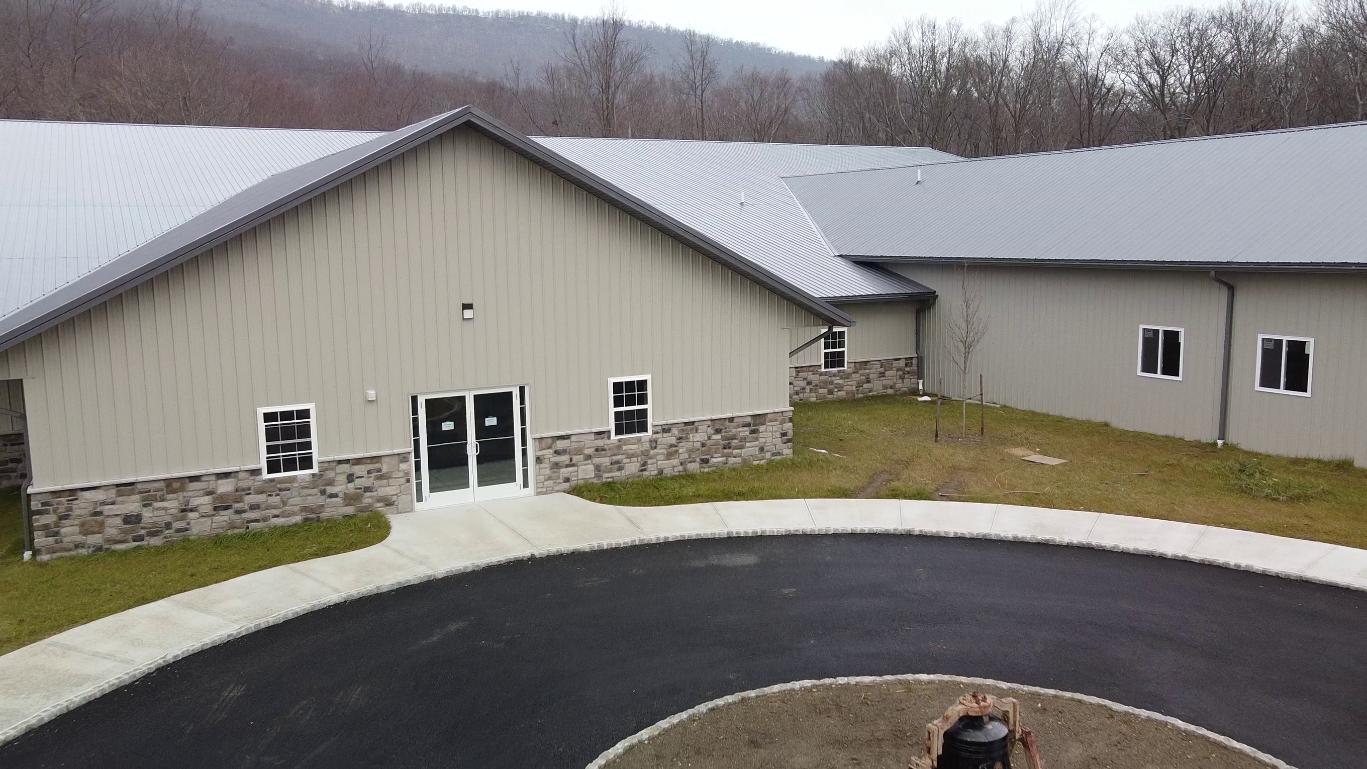Aerial view of a large grey building with stone accents at the base, resembling an elegant arena. The structure, ideal for horse riding events, boasts a metal roof and numerous windows, encircled by a paved driveway and grass. Leafless trees and hills form the scenic backdrop.