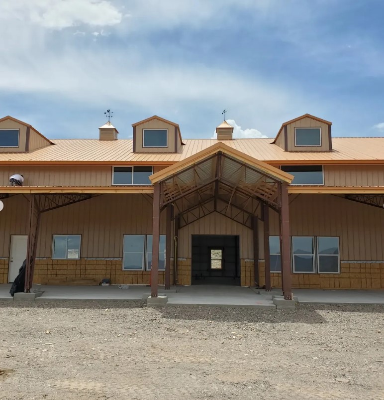 A large beige metal building, reminiscent of a barndominium, boasts an extended entrance and multiple windows set against a clear blue sky. With small upper windows and a covered walkway at the front, the sienna gravel surrounding it adds to the expansive charm.