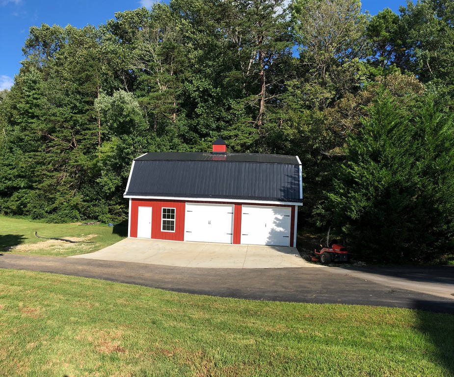 A red and white gambrel-style garage with a black roof sits gracefully in front of a dense forest. A lawnmower rests on the driveway, showcasing its practical charm. The grassy foreground enhances the picturesque scene beneath the clear blue sky.