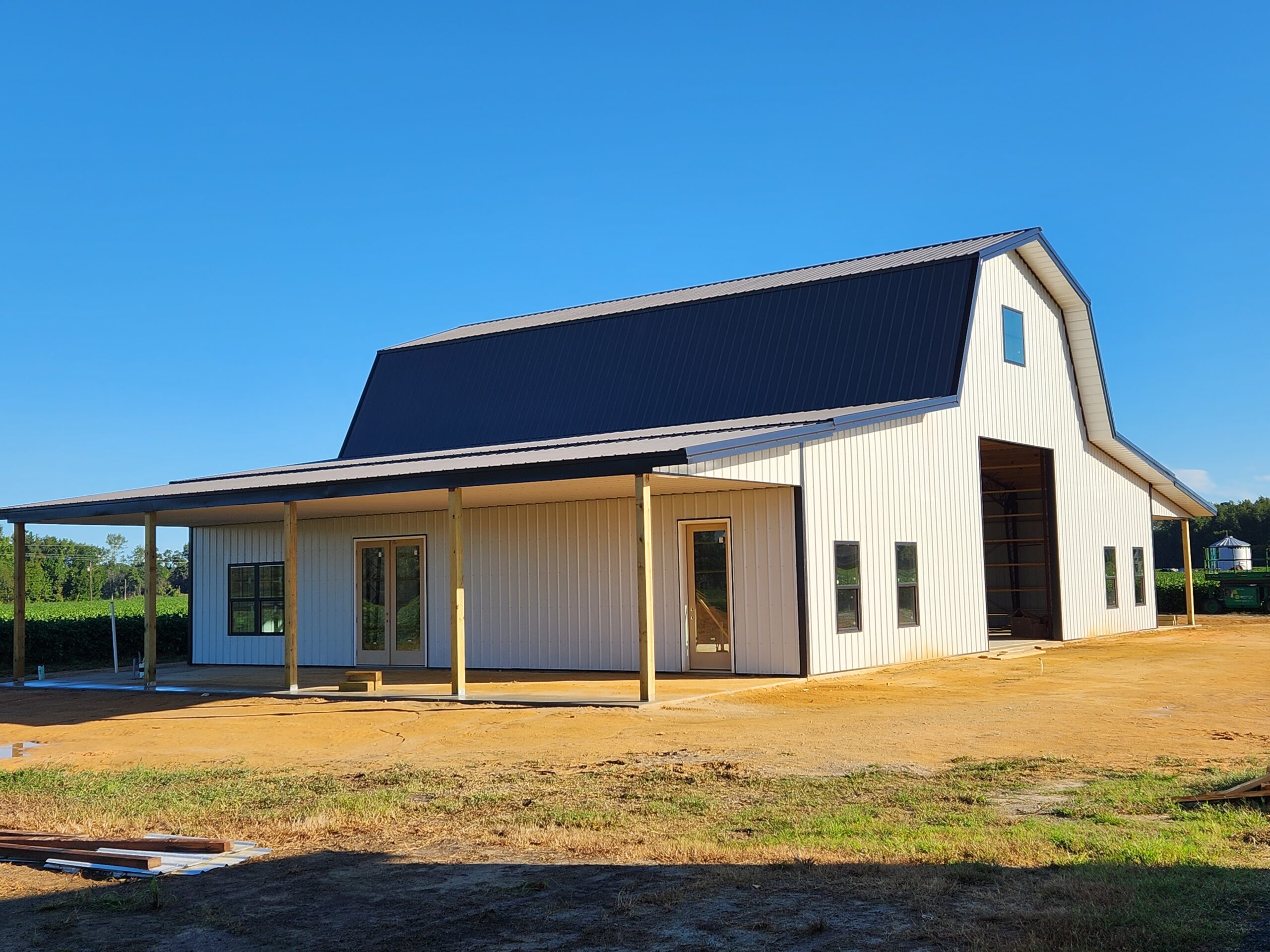A large white metal building with a black roof, resembling a modern garage, stands on a dirt area. It features a covered porch and large sliding doors, surrounded by green foliage under a clear blue sky.