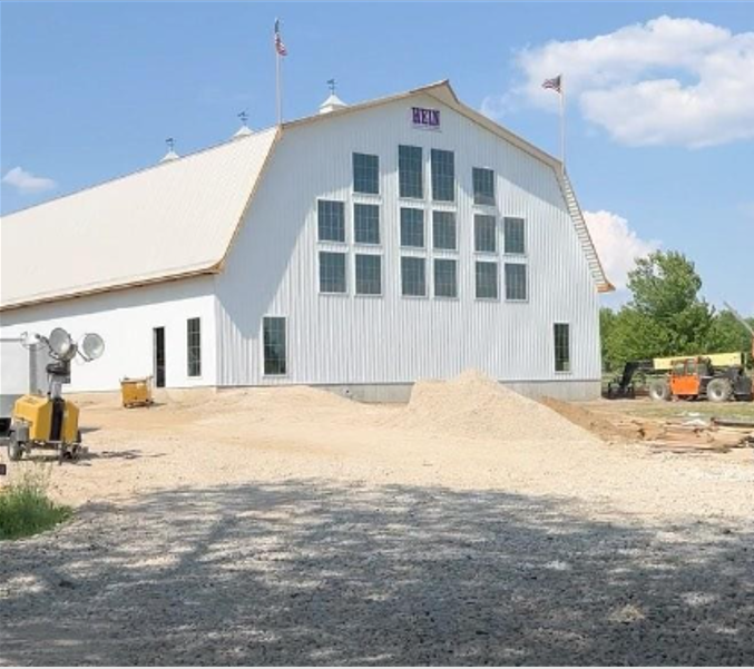 A large white barn with multiple windows in a geometric pattern and flags atop serves as an idyllic wedding venue. With construction equipment and gravel piles in the foreground, the gambrel-roofed structure is framed by trees against a backdrop of blue sky.