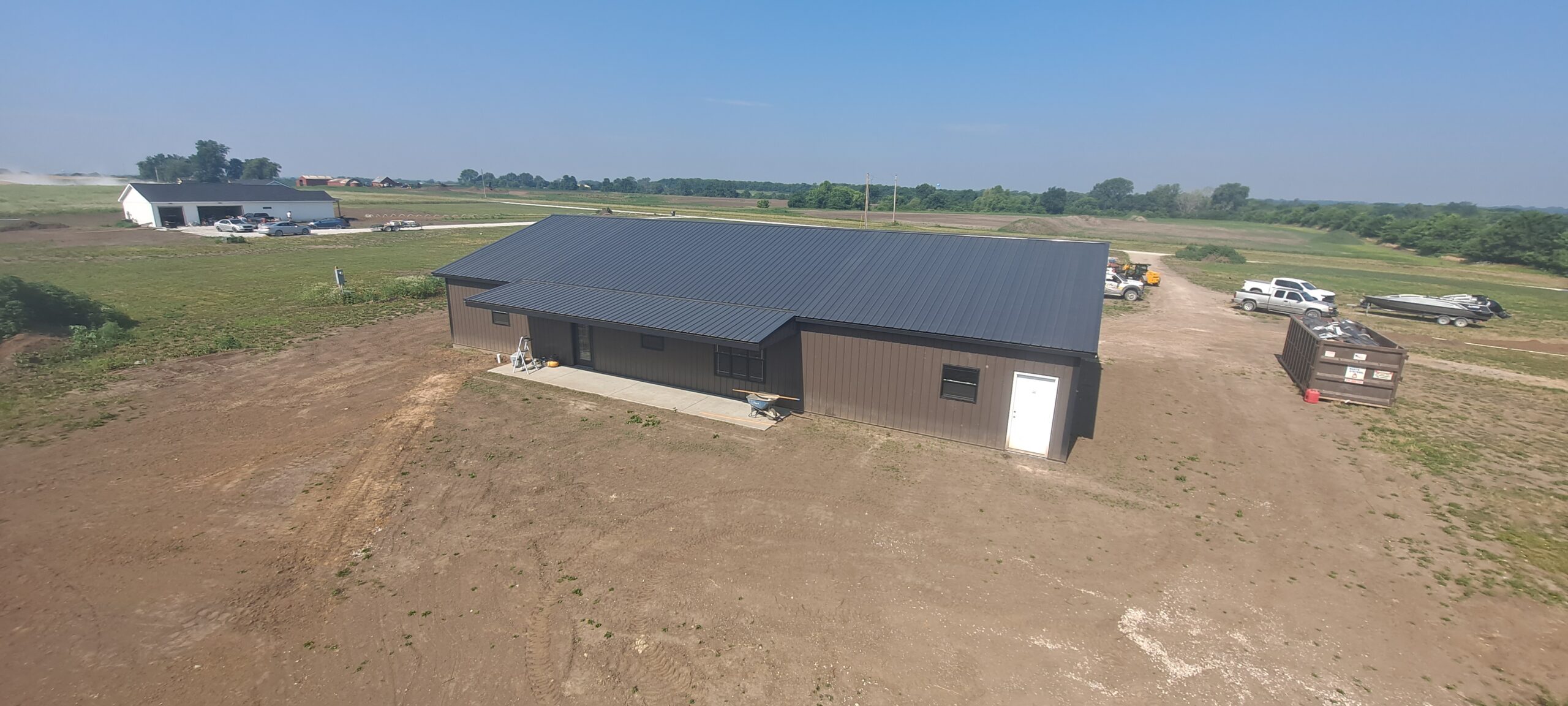 Aerial view of Reece Ranch, revealing a large brown barndo with a sleek black roof in a rural setting. The landscape is mostly dirt and grass, with a few vehicles and a dumpster nearby. Fields and trees stretch into the distance under the clear blue sky.