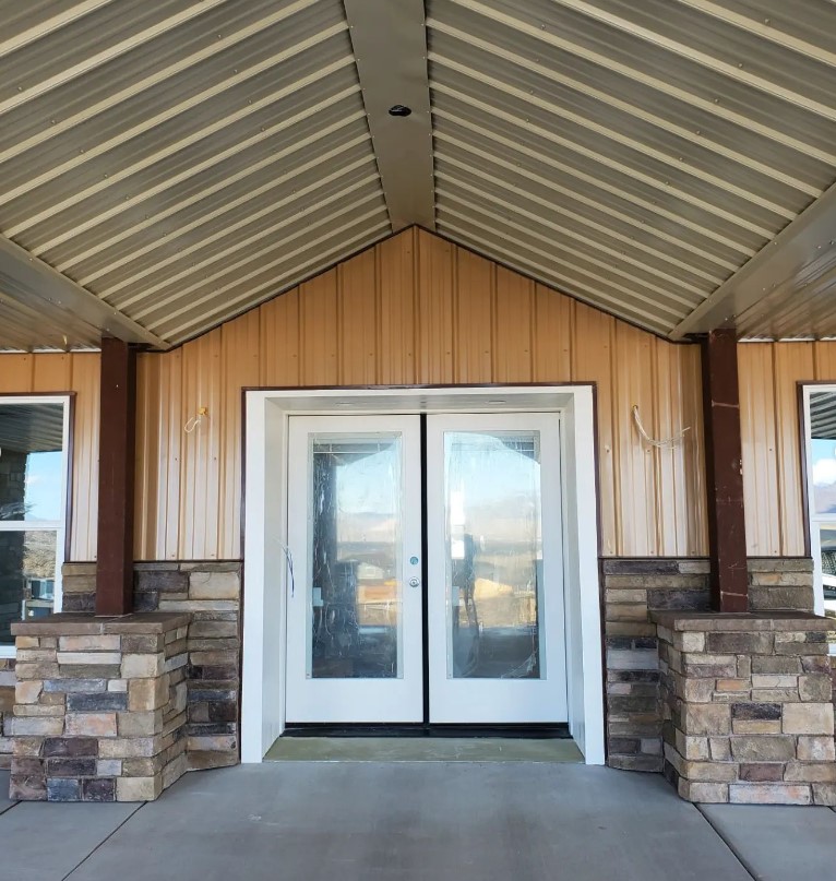 The entrance of this barndominium boasts a sienna metal roof and beige siding. Double glass doors, framed with white trim and flanked by stone pillars, enhance its modern and welcoming appearance.