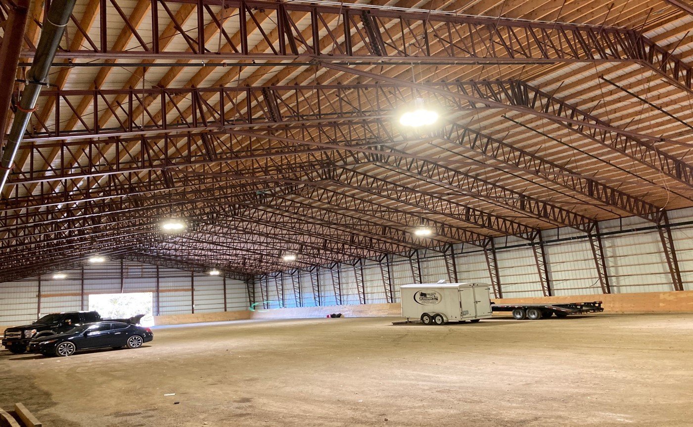 The interior of a spacious warehouse resembles a riding arena with its high, open-beam ceiling. A black car, a white trailer, and a flatbed trailer are parked inside. The corrugated metal walls and gravel-covered ground complete this unique setting.
