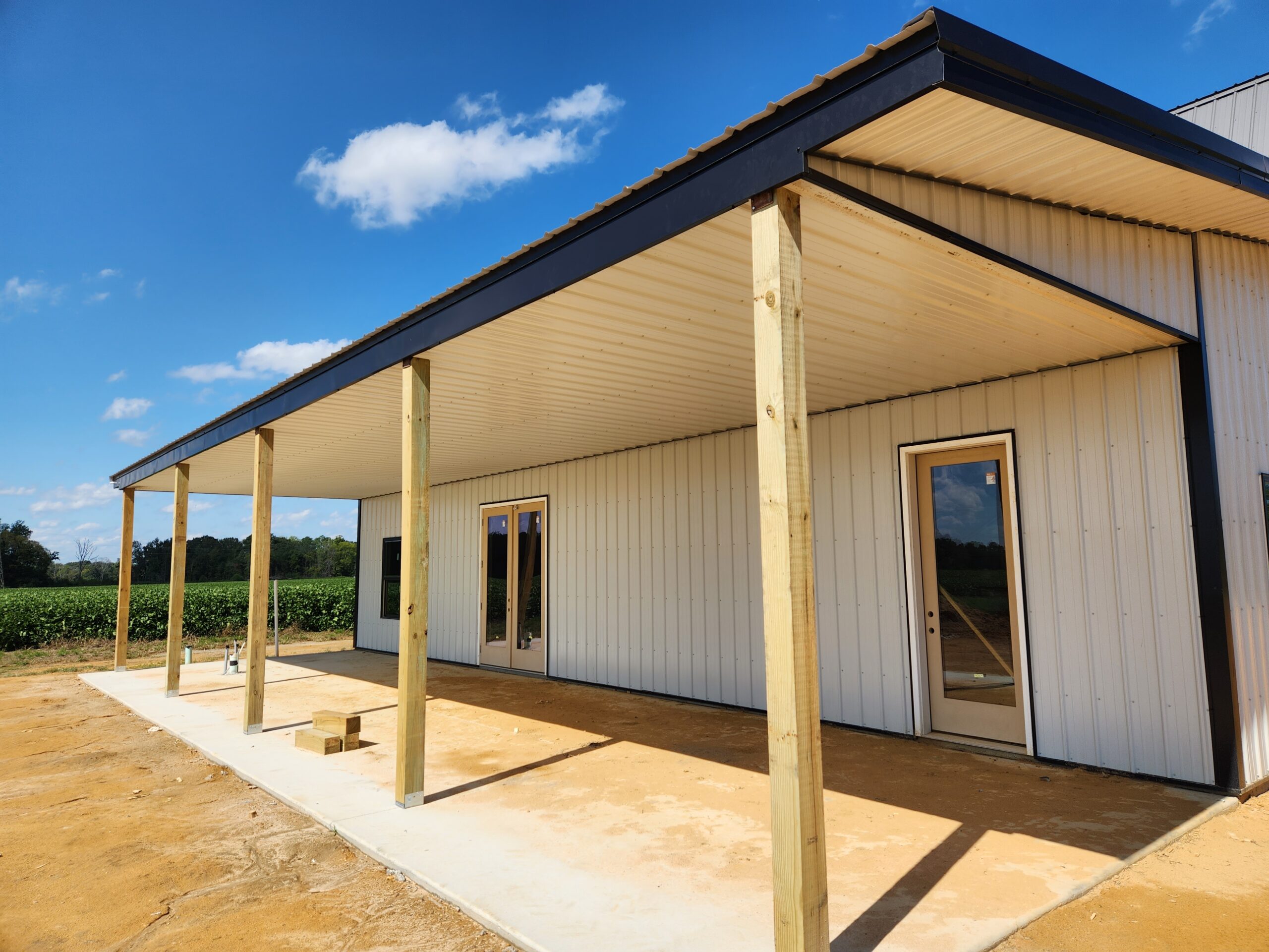 A modern white building with a metal roof and tall wooden pillars supporting a covered porch resembles a gambrel design. The structure is surrounded by a dirt area, with a field and blue sky in the background, adding to its rustic charm.