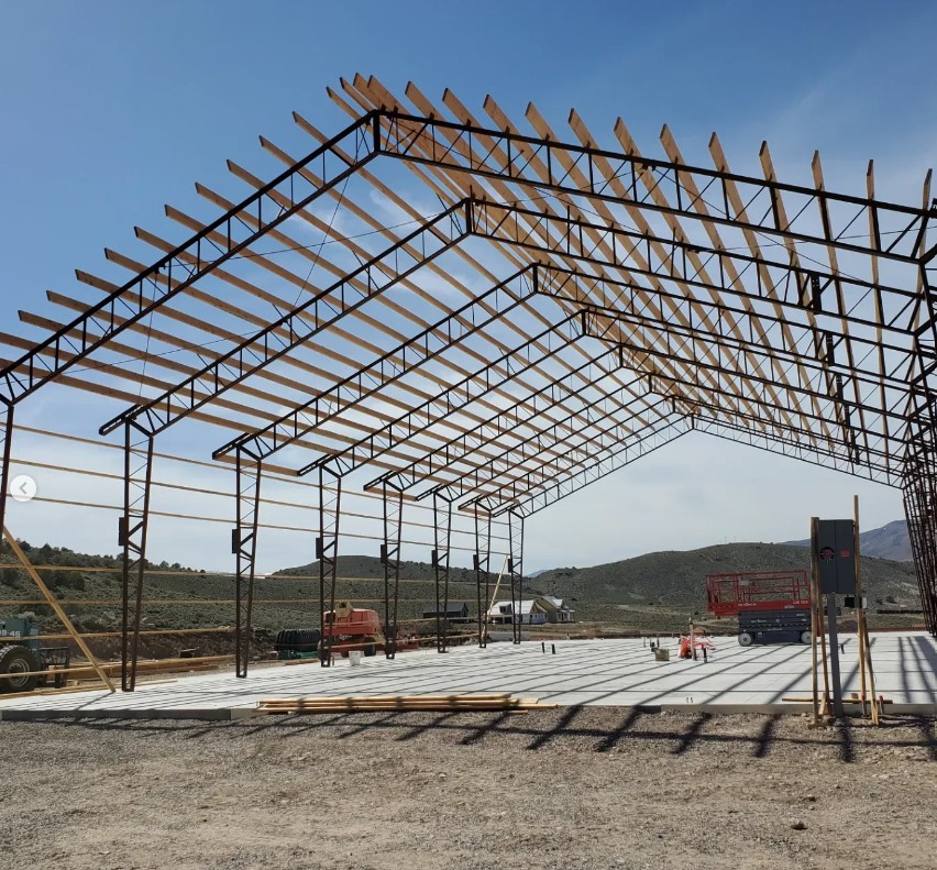 A partially constructed metal frame of a large barndominium stands on a concrete foundation. Sienna wooden beams extend across the top. Several construction vehicles and equipment are on-site under a clear blue sky with mountains in the background.