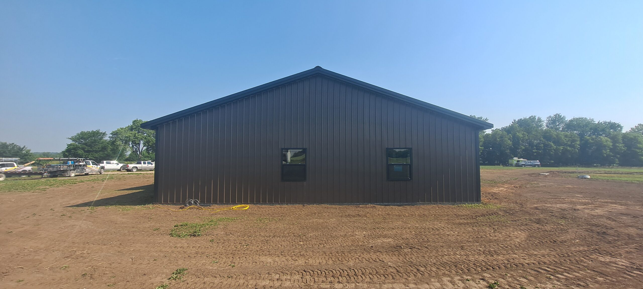 A large, newly constructed dark gray metal building, resembling a barndo, stands on the bare dirt lot of Reece Ranch. The sky is clear and blue, with a few trees and vehicles visible in the background.