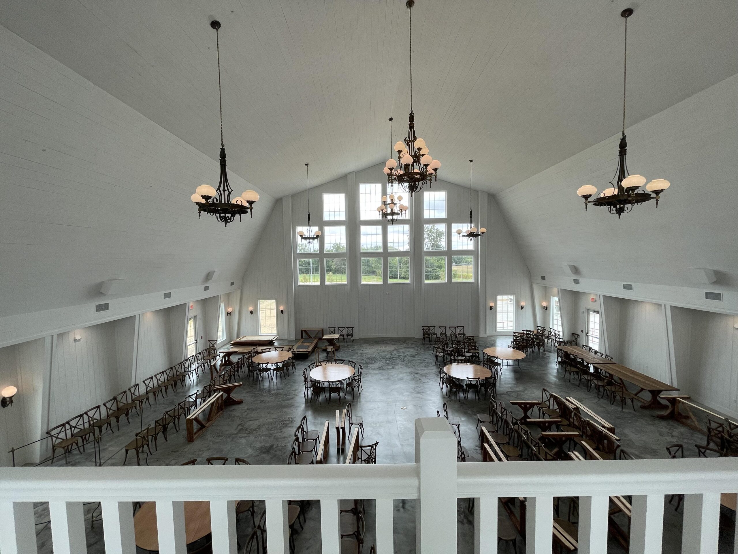 Spacious, well-lit wedding venue featuring a stunning banquet hall with high ceilings and large windows. Several round and rectangular tables with chairs are arranged on the polished floor, while elegant chandeliers hang from the ceiling, providing ambient lighting.