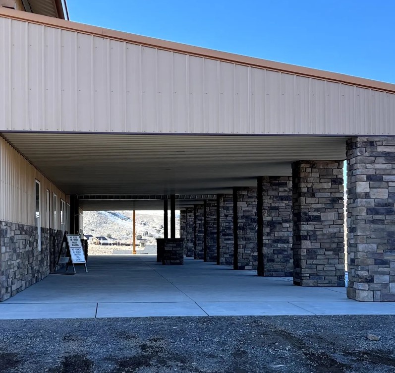 The image showcases a covered outdoor area with stone pillars supporting a metal roof, reminiscent of a rustic barndominium. A sign is visible near the entrance, and a backdrop of sienna-tinted mountains can be seen in the distance under a clear blue sky.
