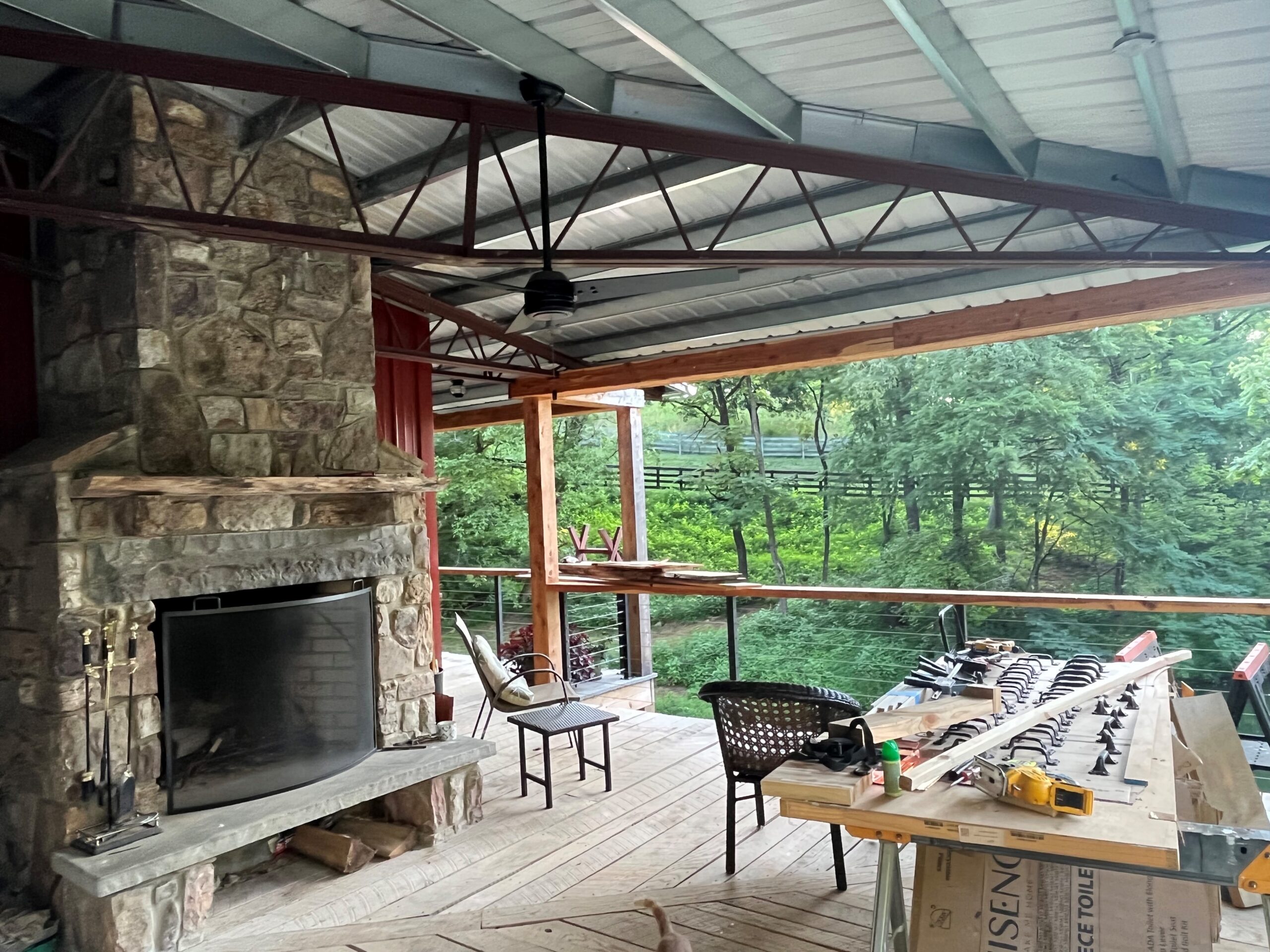 A spacious covered patio with a stone fireplace and ceiling fan adds charm to this asymmetrical lakehouse. The wooden deck overlooks lush greenery, while patio furniture and a table with tools suggest ongoing construction or renovation work, promising future improvements.